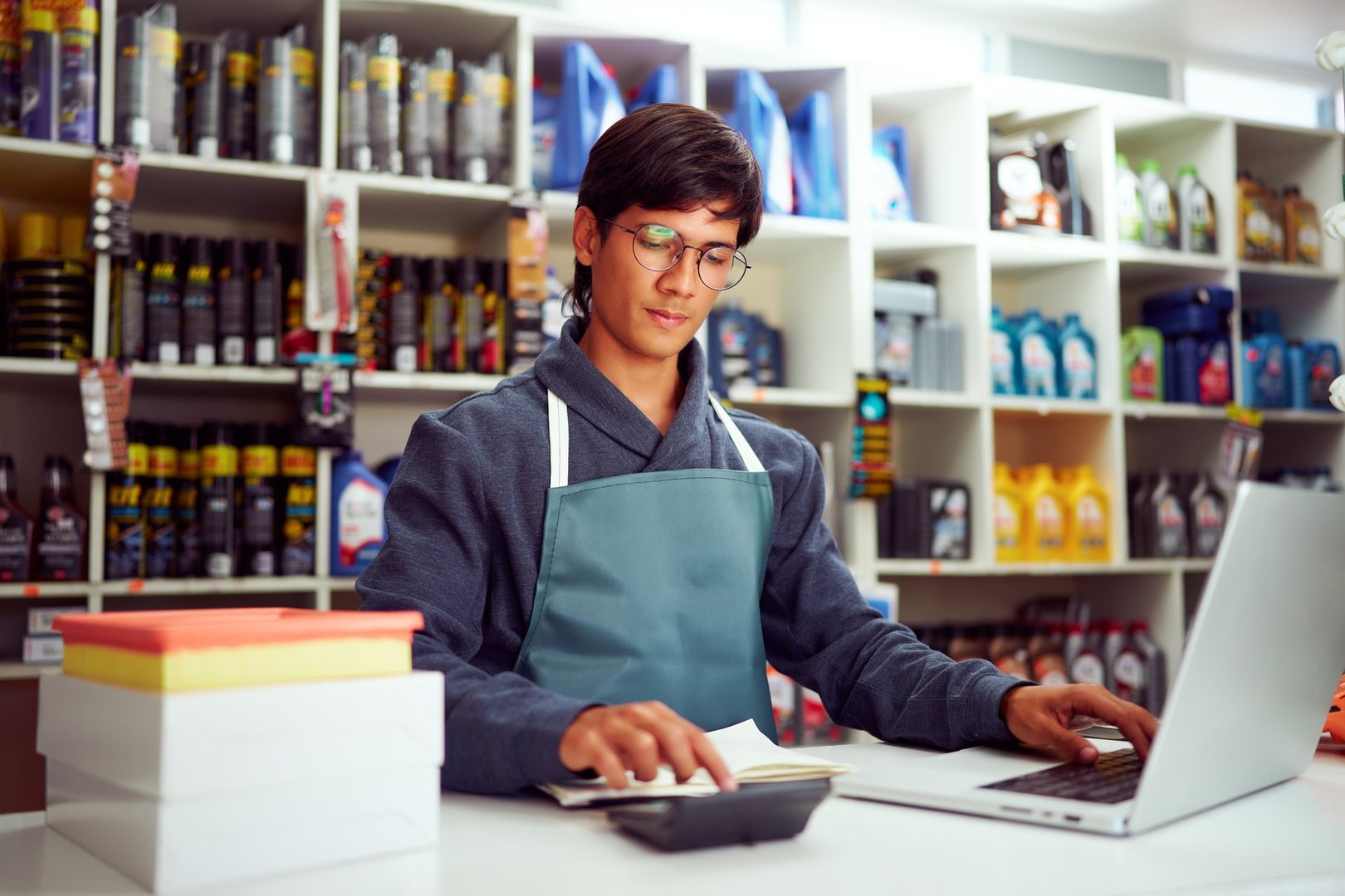 Young male shopkeeper using calculator and laptop while working in his store