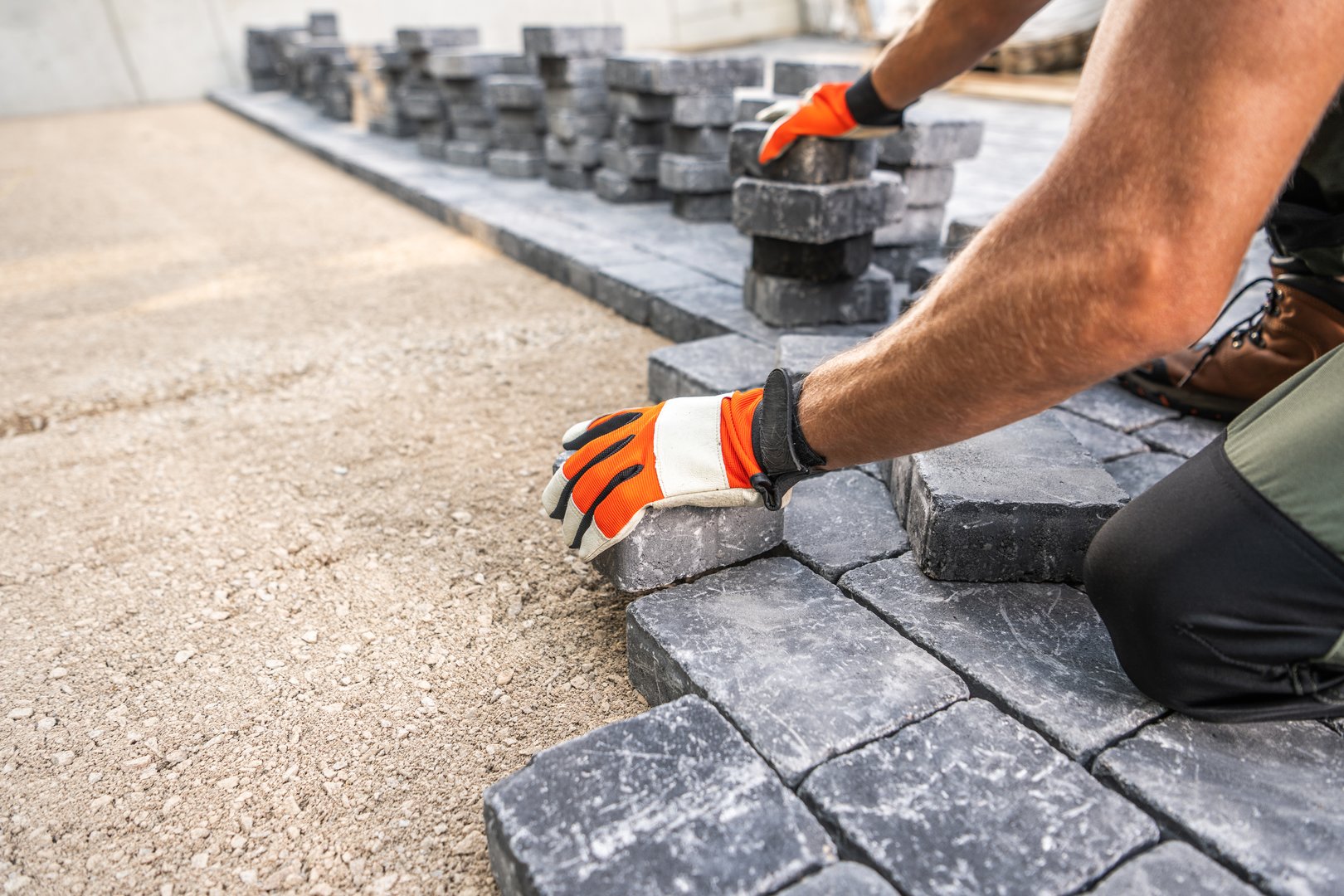 A worker is carefully placing paving stones on a prepared surface at a construction site. The person is dressed in safety gear and focused on aligning the stones properly while laying them in a structured pattern.
