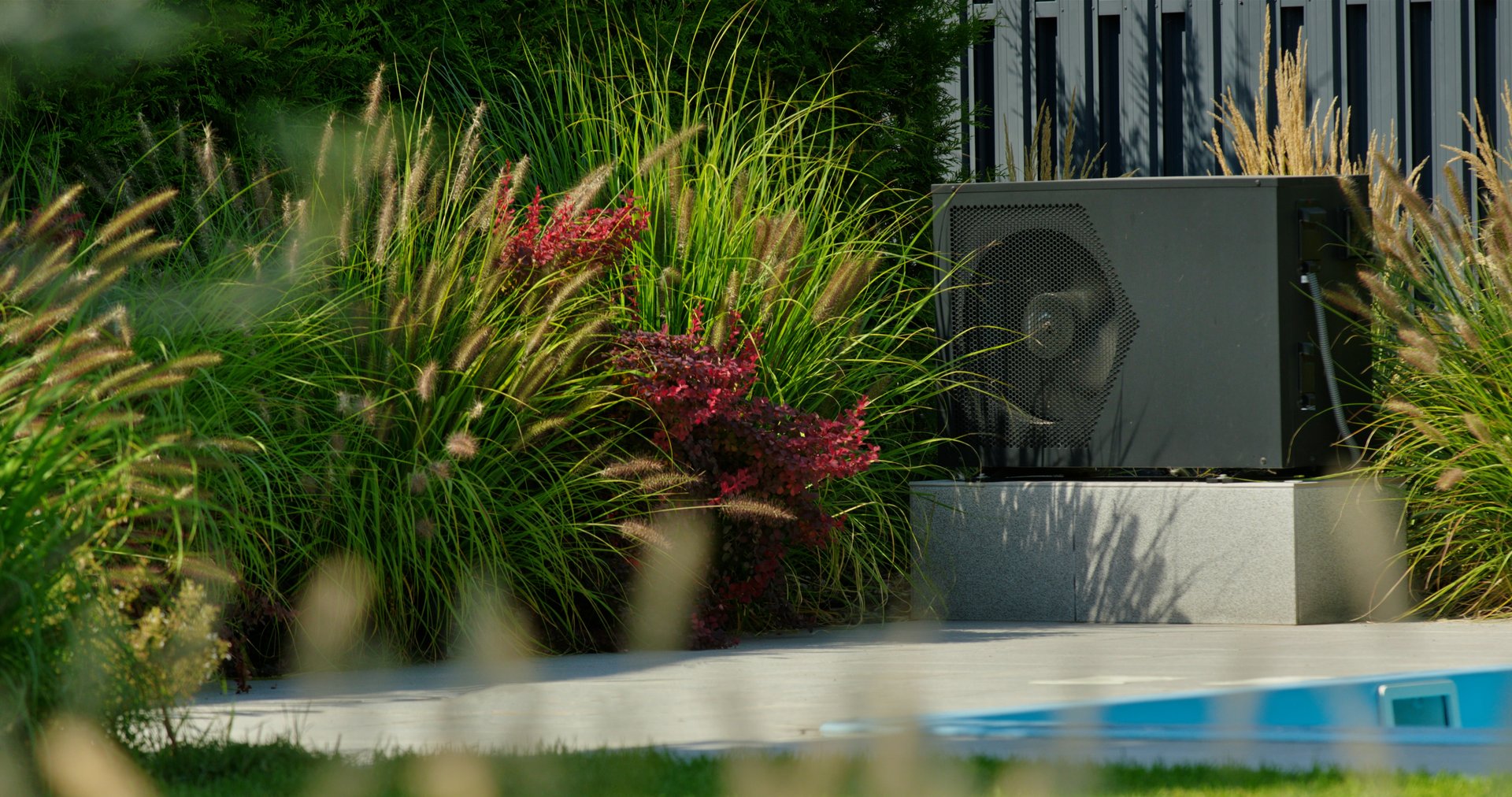 An outdoor module of a heat pump, strategically placed near a backyard pool, nestled among lush, blooming plants. The setup combines modern technology with a natural landscape, enhancing the aesthetic appeal and energy efficiency of the home.