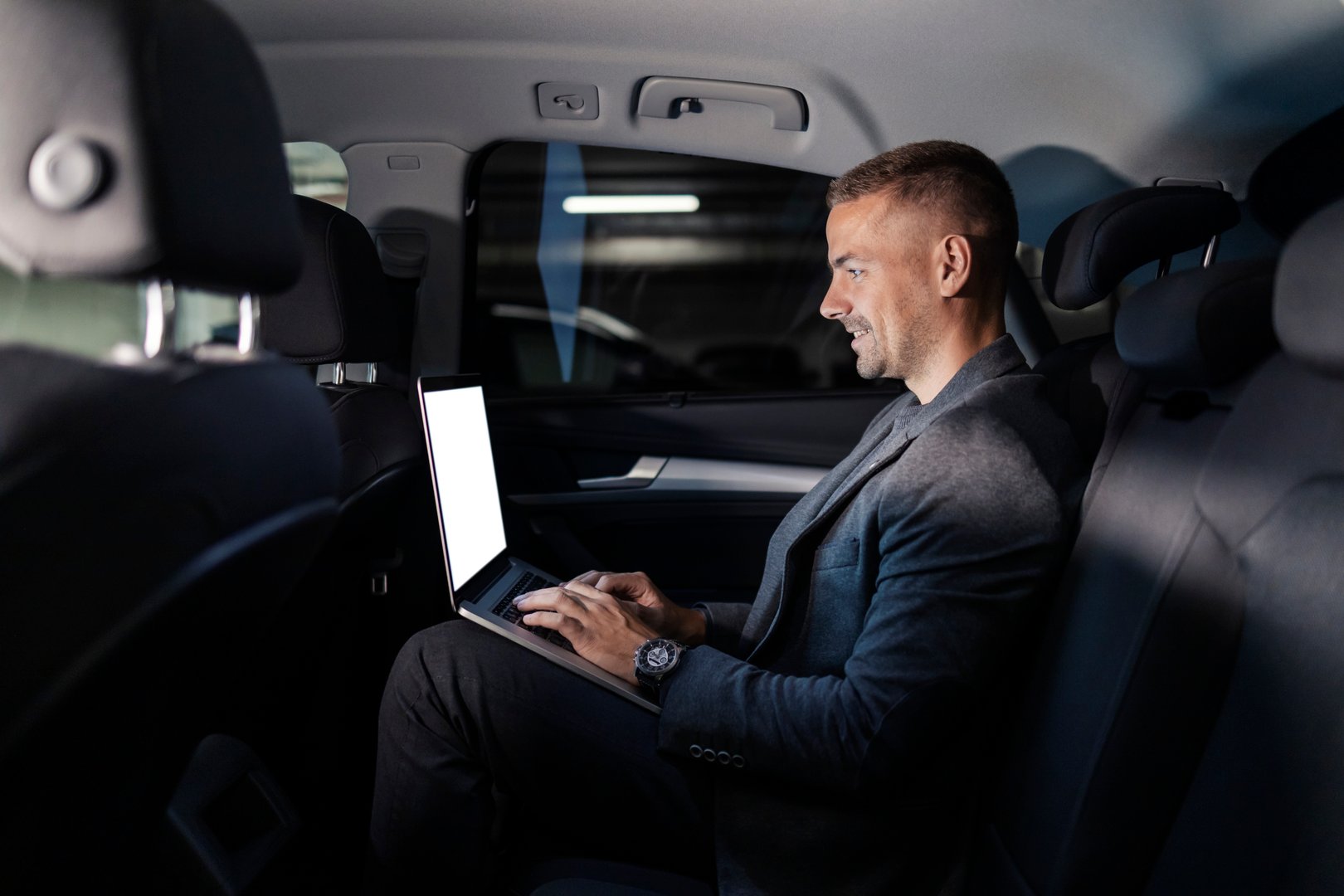 Profile of a young occupied businessman sitting on backseat and working on his laptop.
