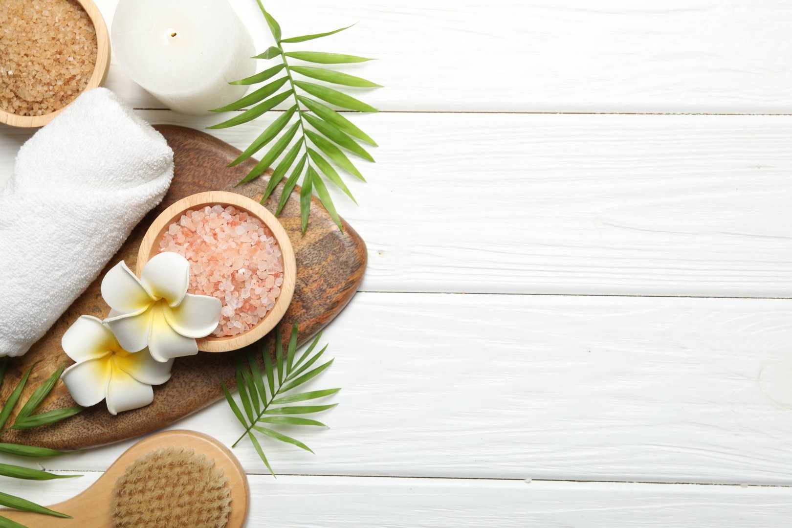 Spa composition with sea salt, plumeria flowers and burning candle on white wooden table, flat lay. Space for text