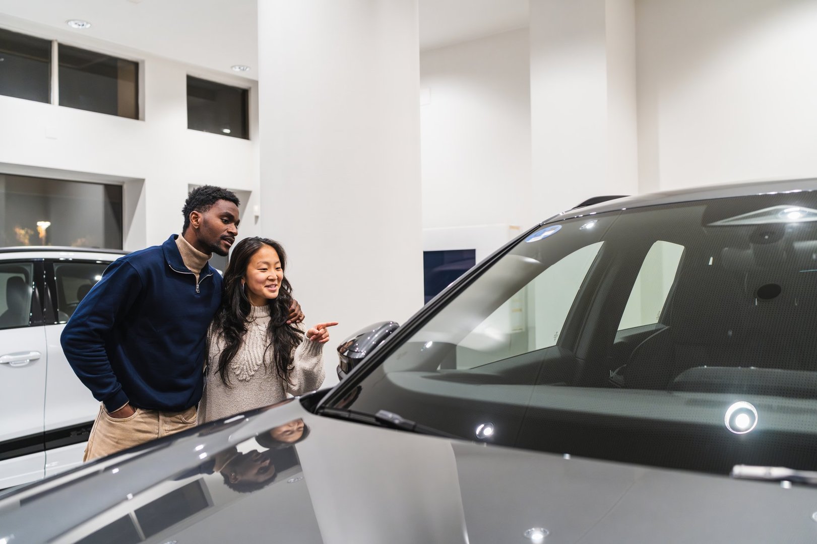 Young multi-ethnic couple choosing new car in dealership showroom, hugging and pointing at vehicle