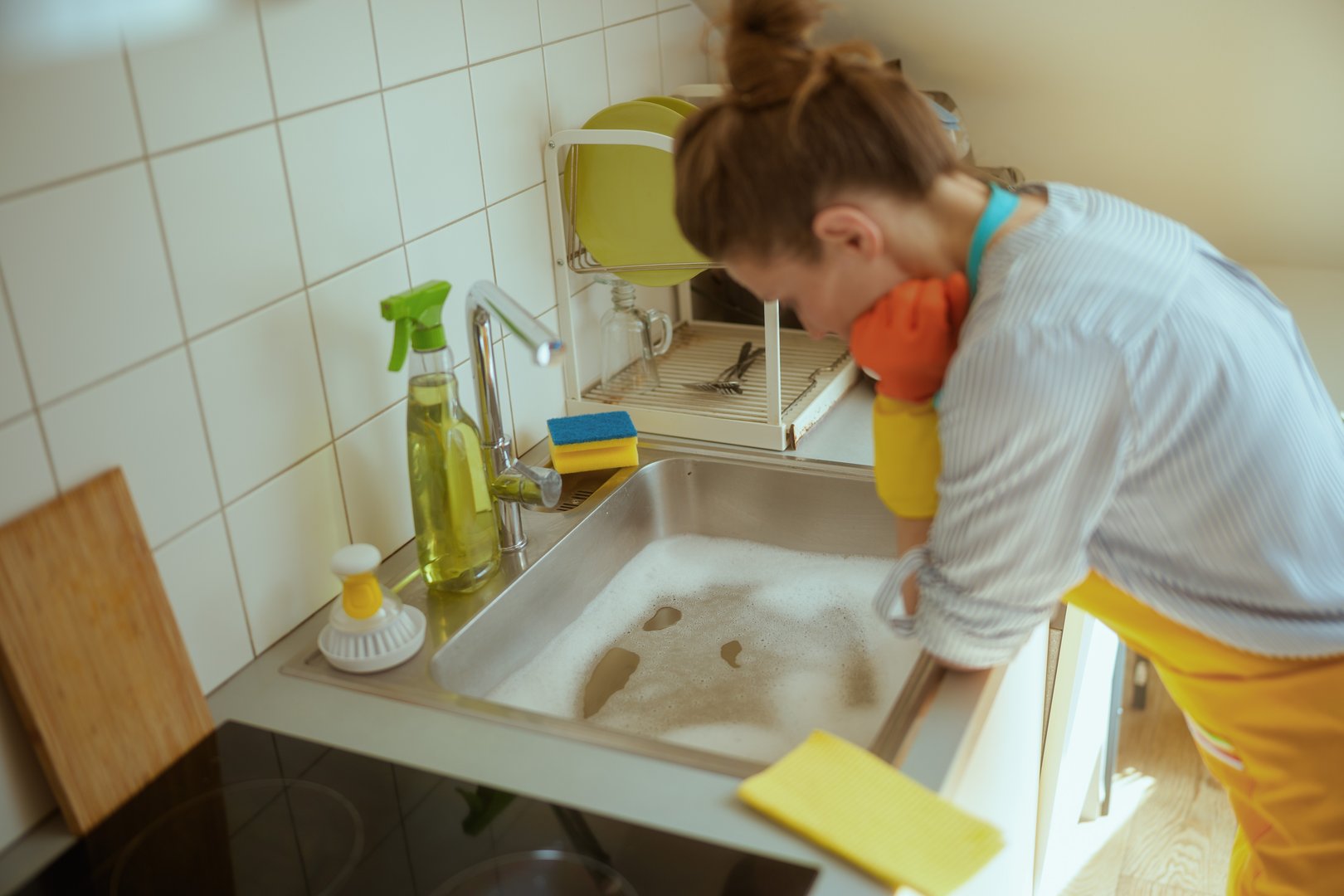 Wearing gloves and apron, a frustrated woman leans toward a full soapy sink that wonât drain, overwhelmed by a kitchen pipe problem.