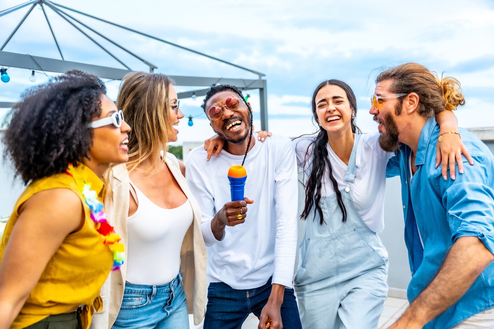 Five cheerful multi-ethnic friends singing karaoke, enjoying laughter and connection during a vibrant rooftop party filled with joy
