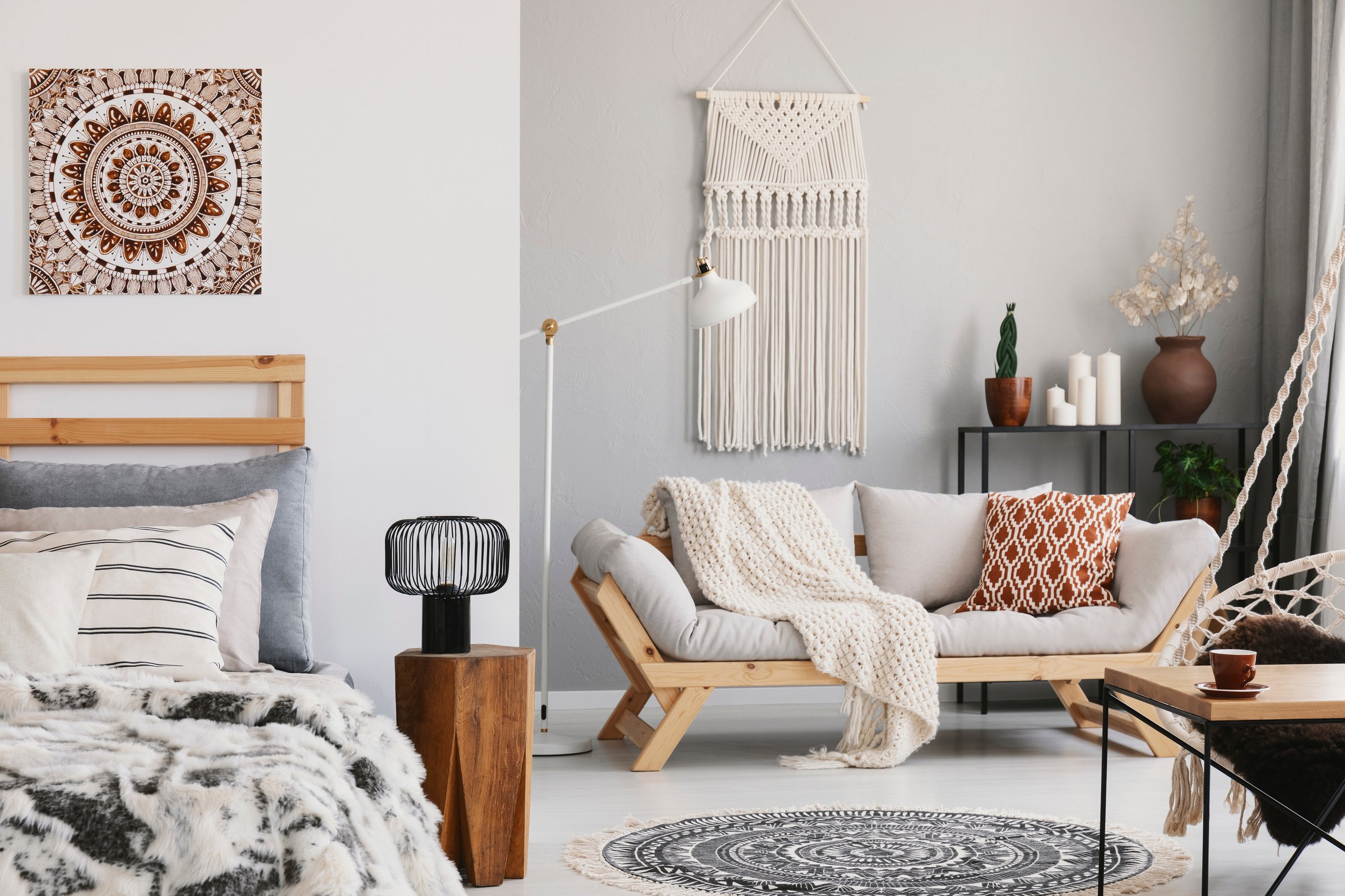 Small open space flat interior with beige sofa with cushion, macrame on the wall, rack with candles and plants and bed with pillows