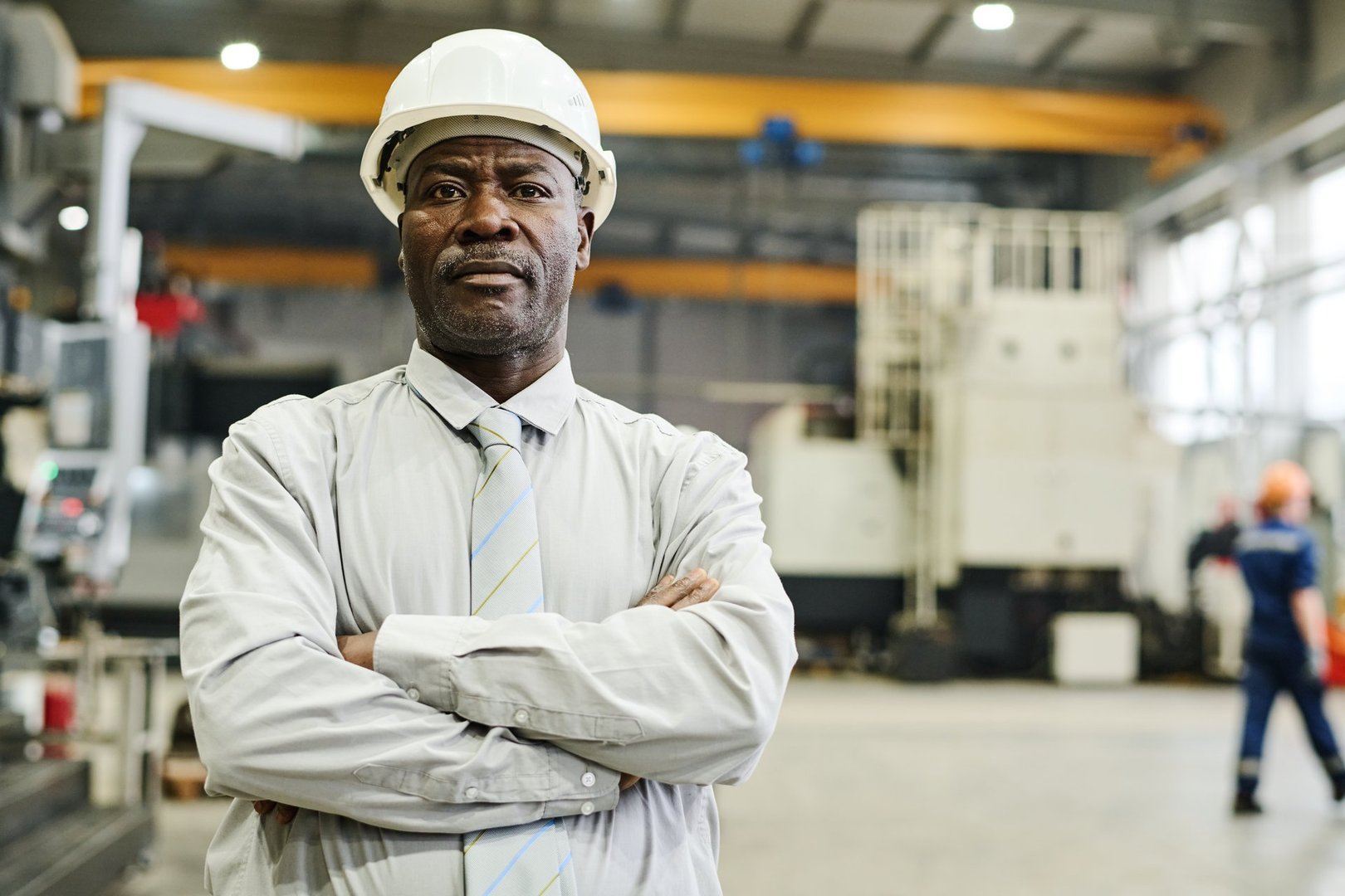 Portrait of African American engineer wearing white hardhat standing in factory with arms crossed, background showing industrial equipment and workers