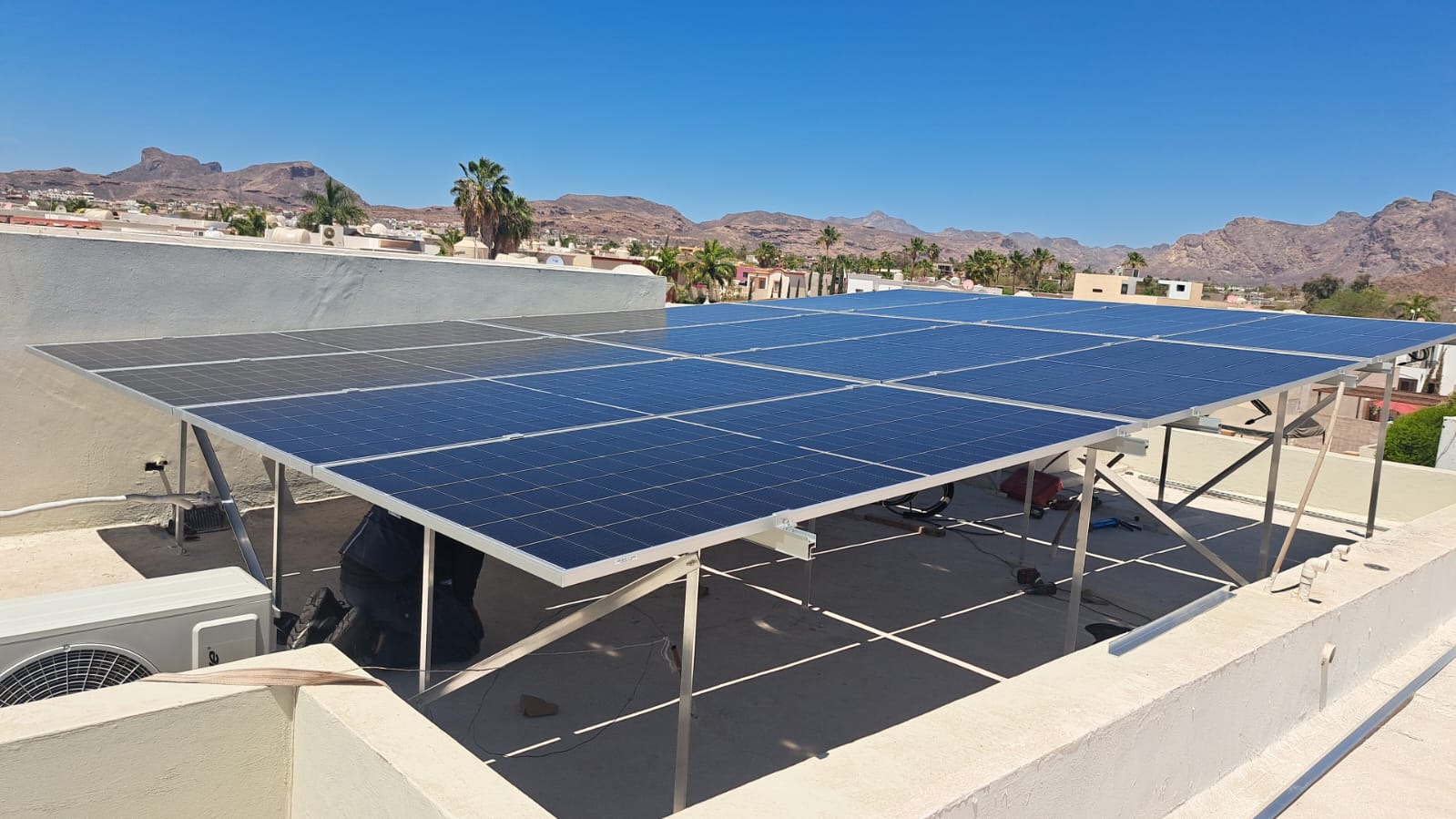 Solar panels installed on a flat rooftop with a view of desert mountains and palm trees in the background under a clear blue sky.