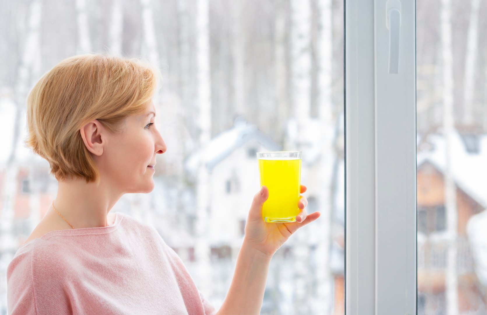 A nice smiling woman in a pink sweater looks at a glass of yellow vitamin drink in her hand, standing by the window in winter