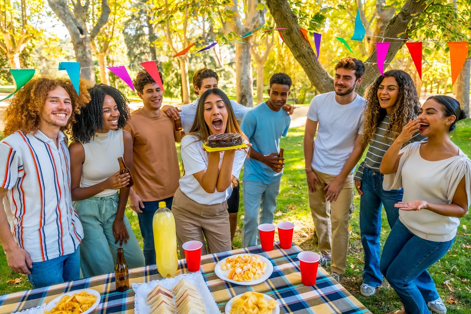 Group of cheerful multi-ethnic friends enjoying a birthday party with cake, snacks and drinks, having fun together outdoors