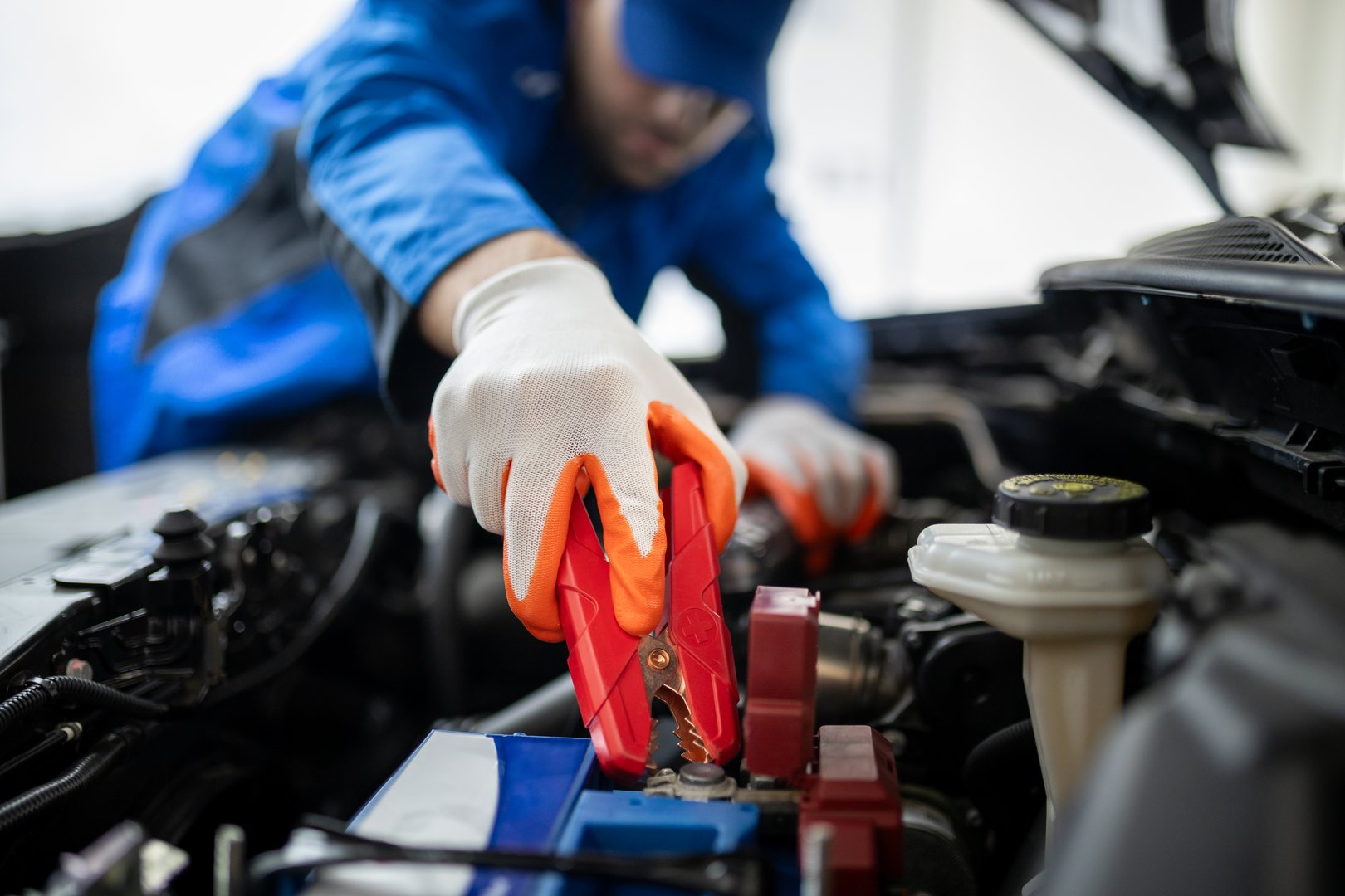A mechanic in a blue uniform is using jumper cables to connect a car battery in a repair shop. The focus is on the connection process, highlighting the tools used.