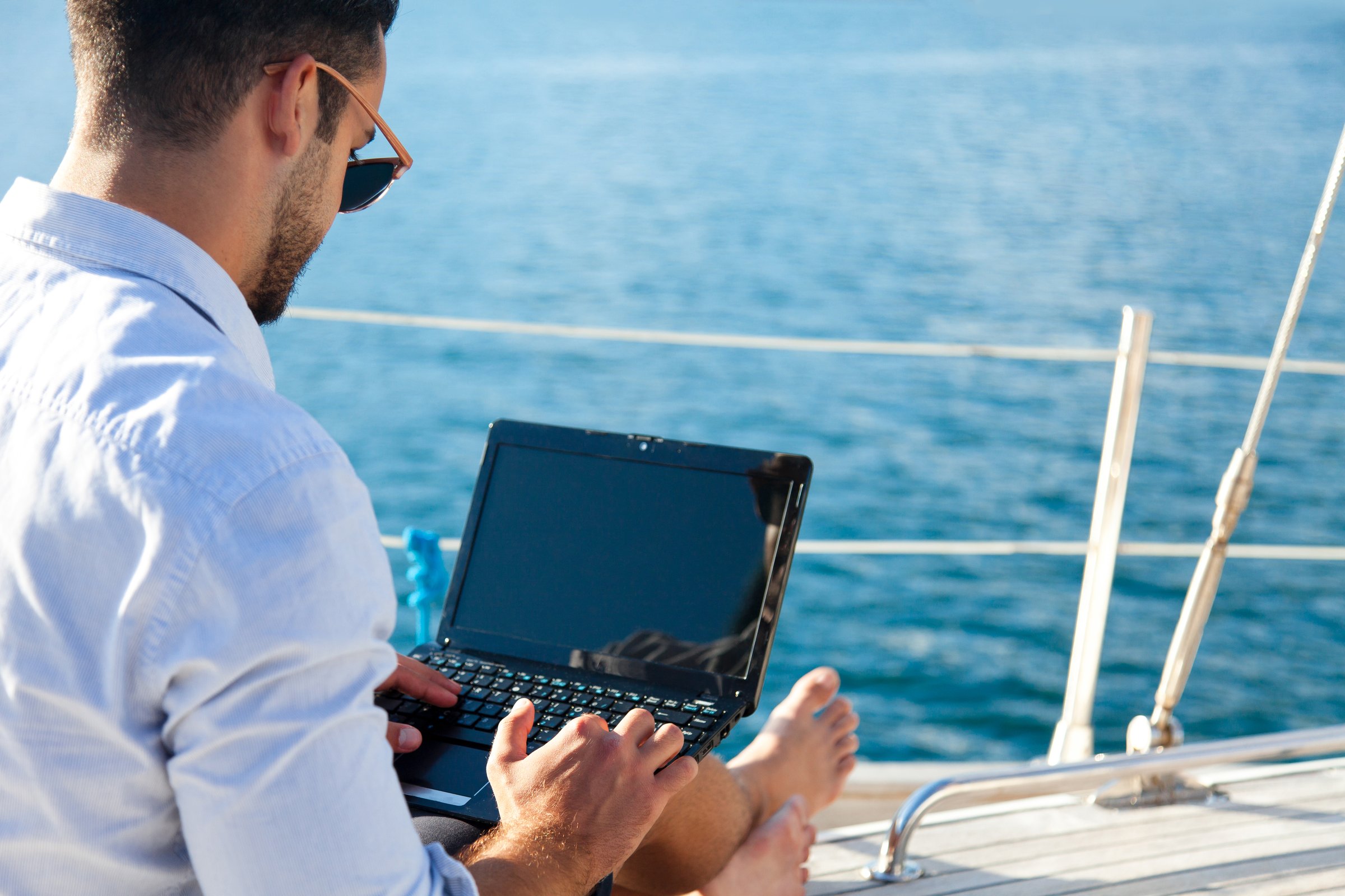 Man working on yacht by sea with laptop. Traveling on sailboat. Traveler using computer, Internet. Freelancer workplace and office in vacation. Successful business lifestyle. Close-up of blank screen.