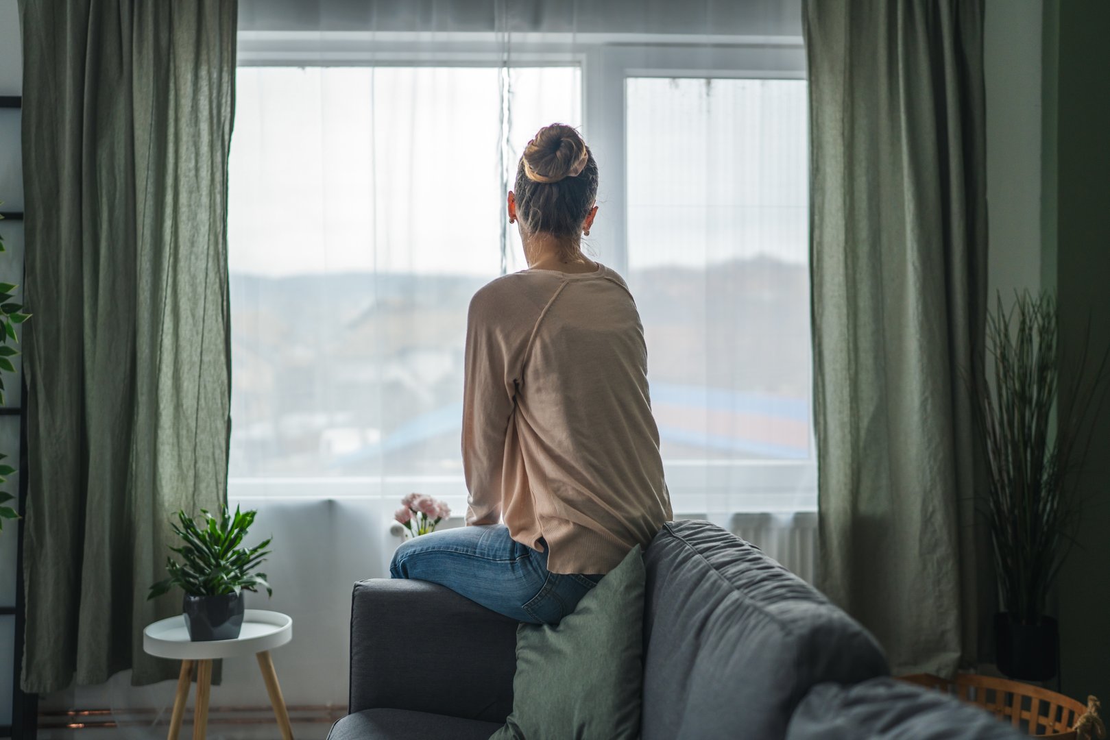Woman sitting on a sofa, back to camera, gazing out a sunlit window in quiet contemplation