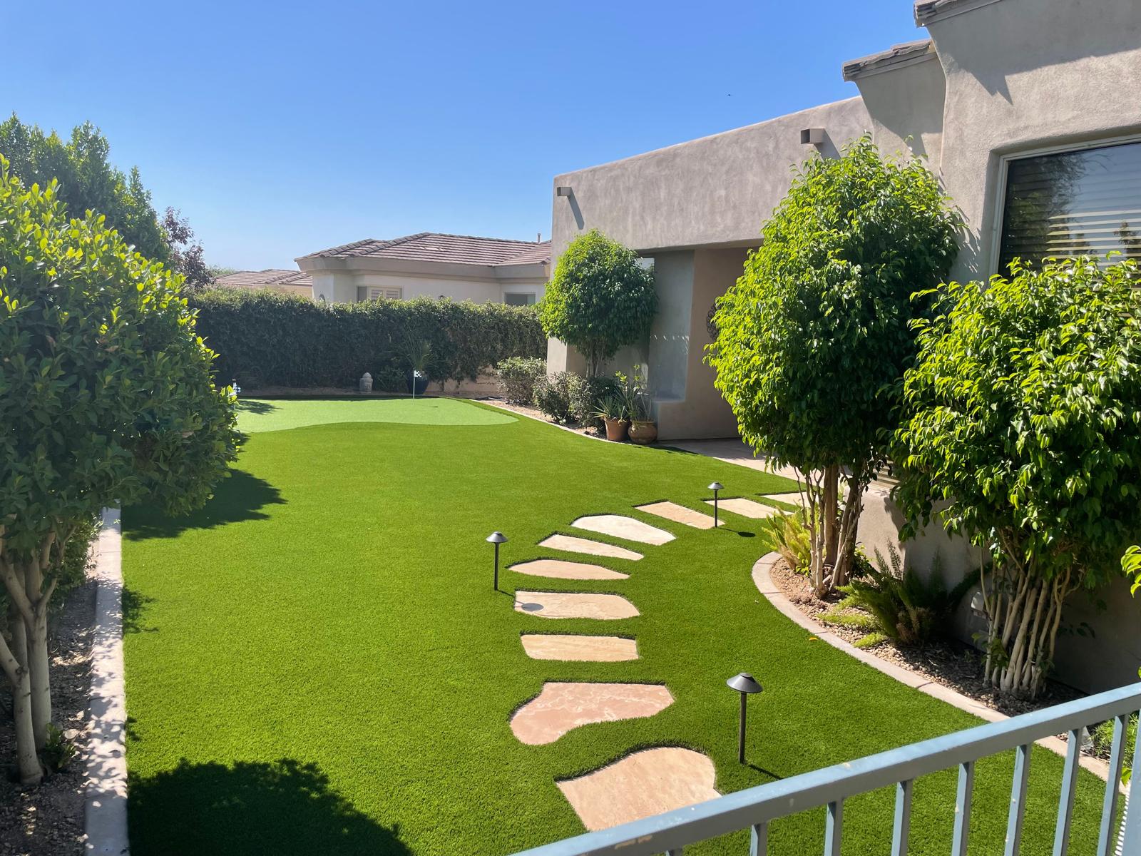 Well-maintained backyard with artificial grass, stepping stones, potted plants, and neatly trimmed bushes under a clear sky.