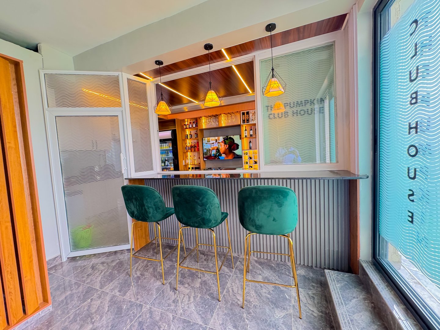 Modern home bar with green velvet stools, pendant lights, and a stocked shelving unit with liquor bottles.