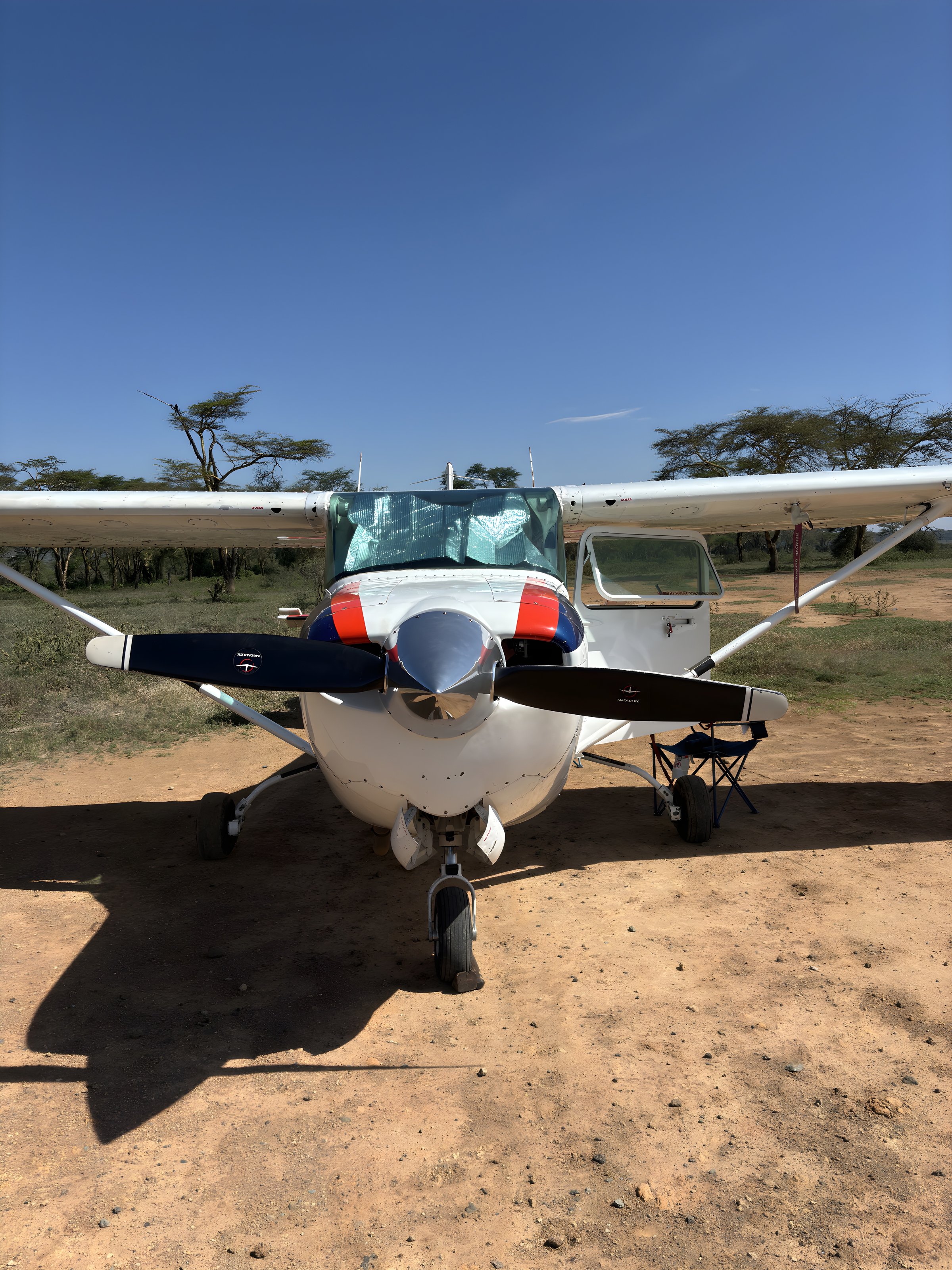 Small white aircraft parked on dirt with trees and clear blue sky in the background.