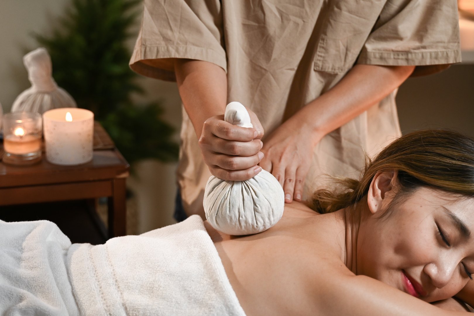 Lady receiving hot herbal ball spa massage, masseur gently compresses herb bags on woman back in warm lighting of candles at spa salon.