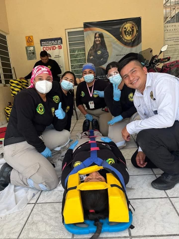 Group of medical responders posing with a person secured on a stretcher, featuring COVID-19 protocols banner in the background.