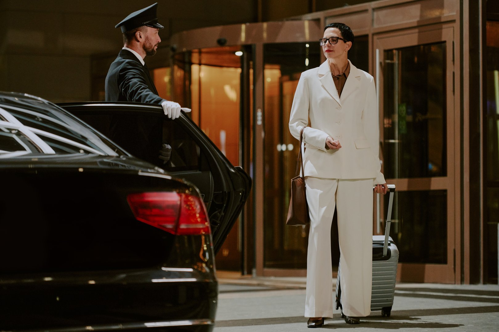 Caucasian middle aged woman standing outside hotel entrance holding suitcase and handbag while Black man chauffeur in uniform opening car door, both engaging in business travel scenario