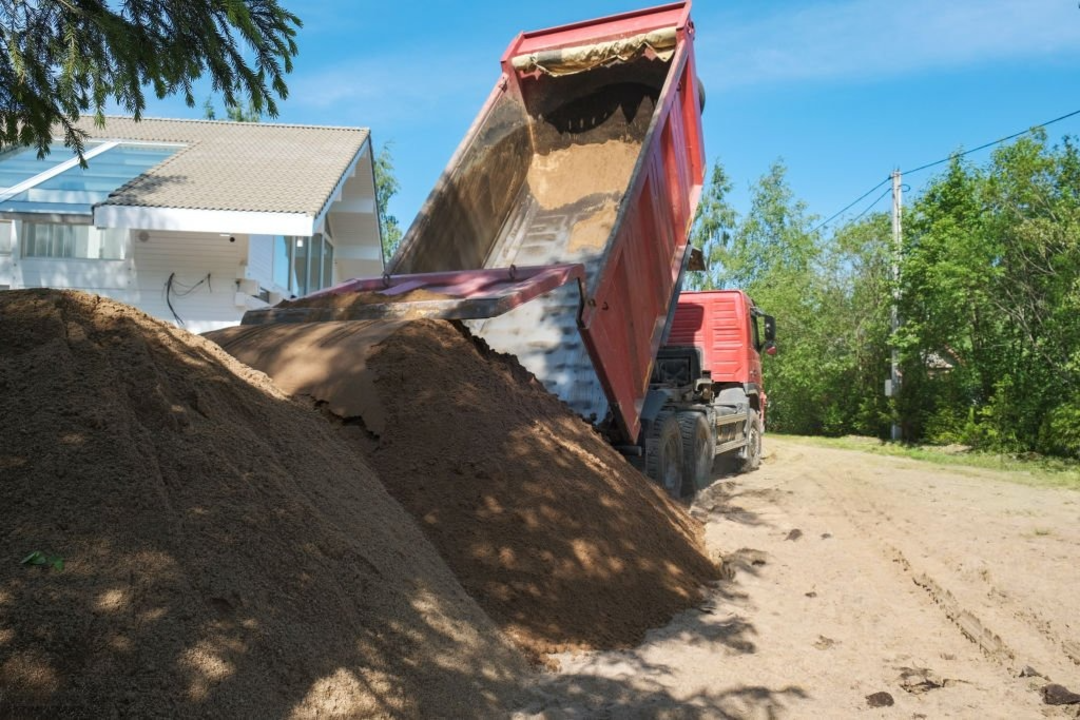 A red dump truck unloading sand onto a residential property with a white house and trees in the background.