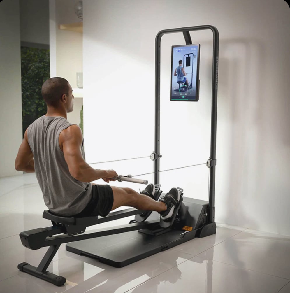 A man exercises on a rowing machine equipped with a digital display showing a trainer, in a modern indoor setting.