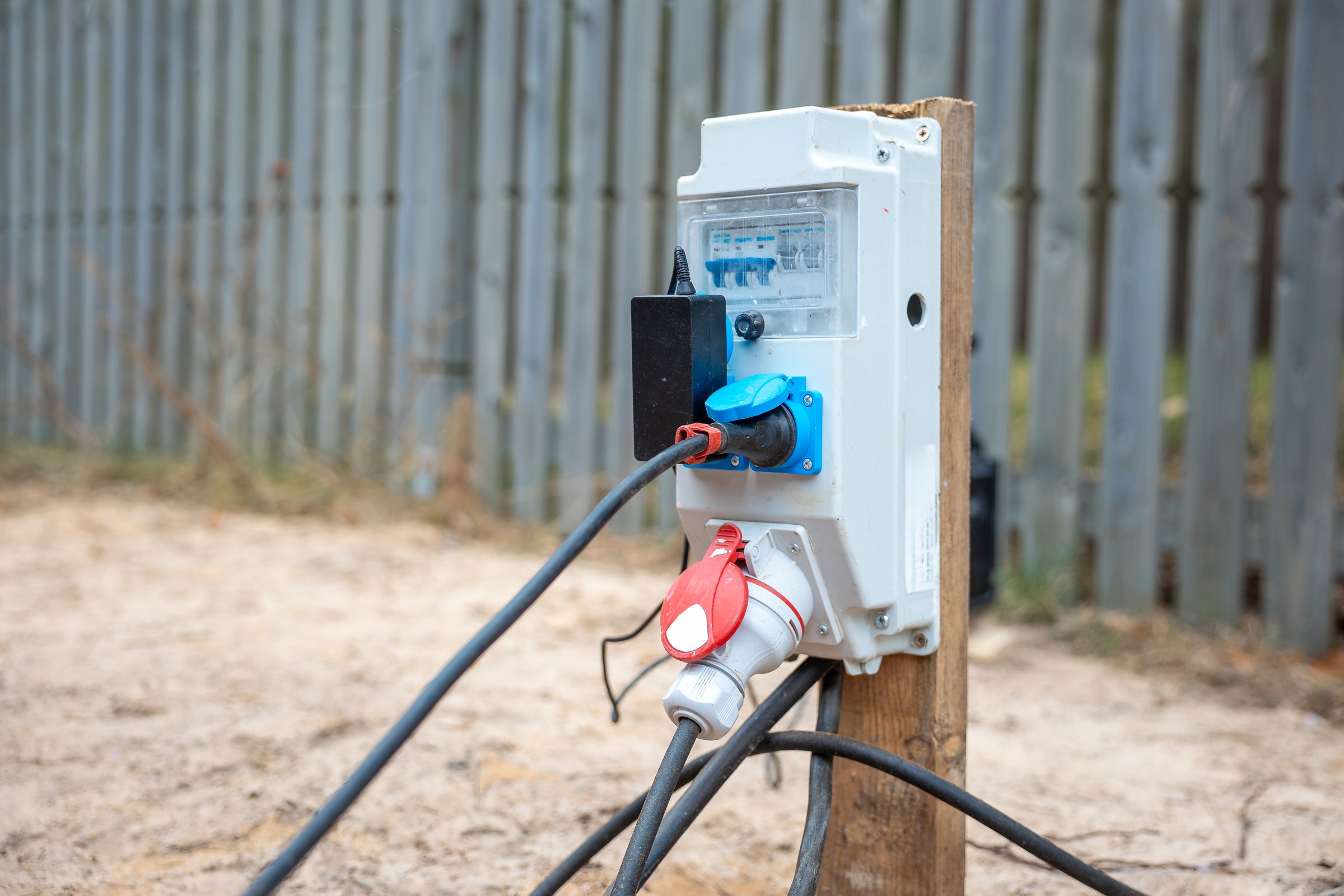Outdoor electrical distribution box on wooden post with transparent cover, circuit breakers, red and blue plugs, and cables at construction site.