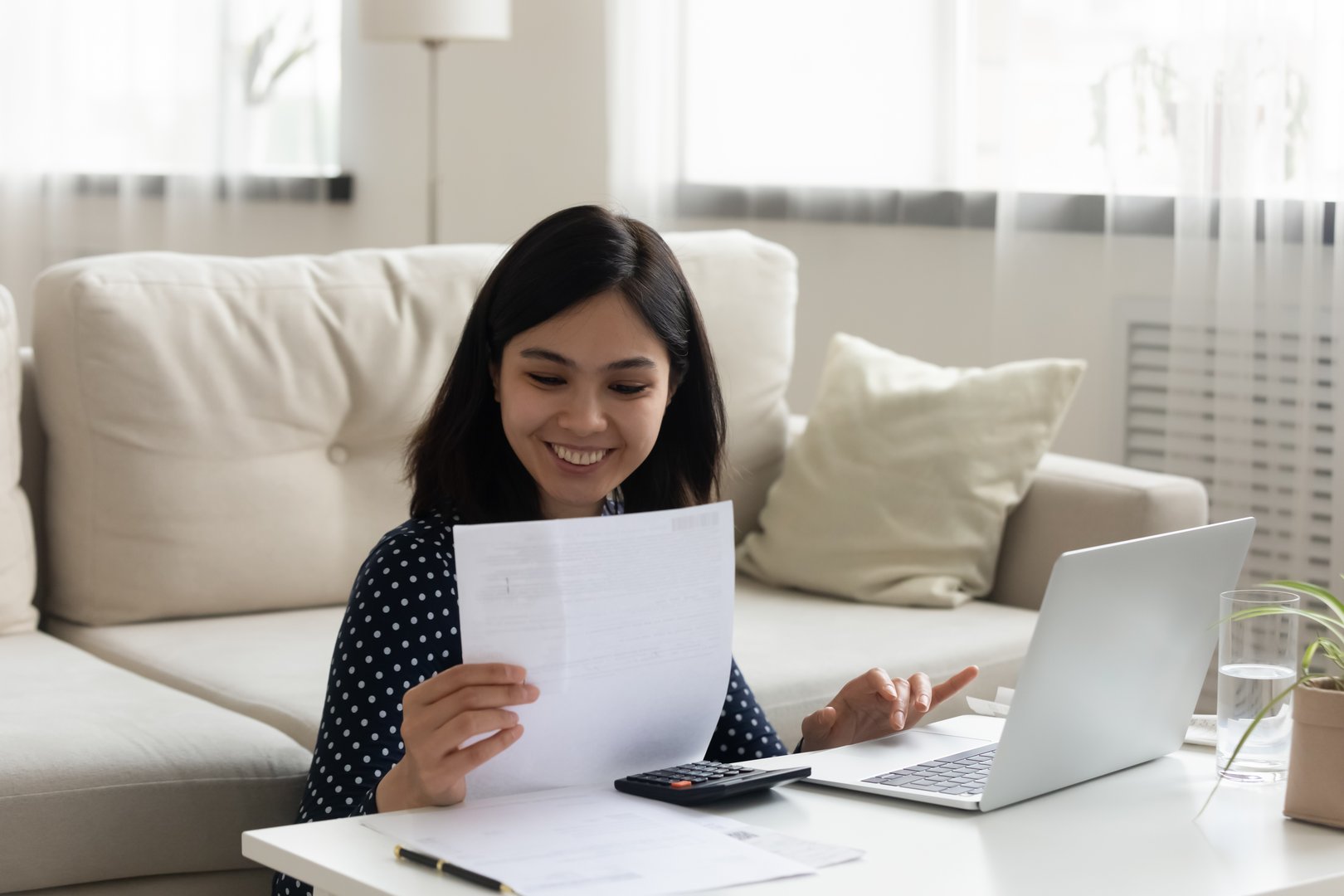 Confident asian female read paper bill invoice prepare to make payment using computer app. Young vietnamese lady engaged in paperwork at home office desk review loan mortgage document check sum online