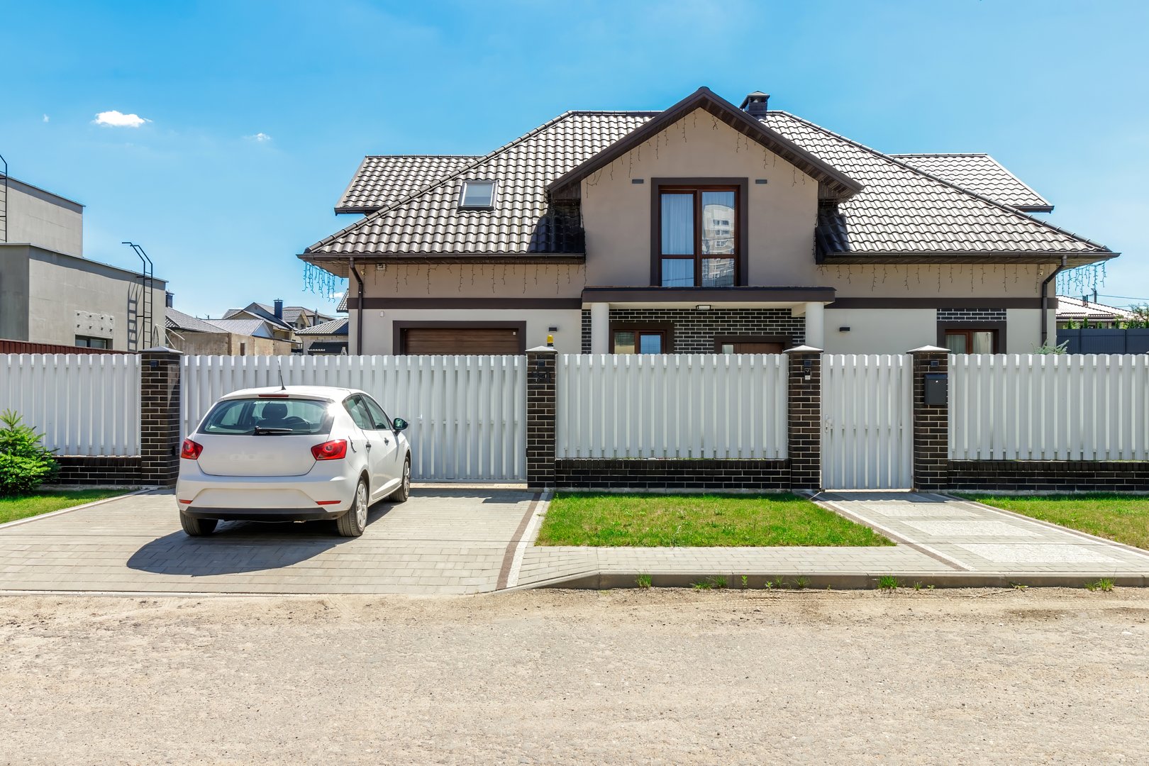 A modern suburban home featuring a driveway with a parked car, white fence, brick details, and a clear sunny sky.