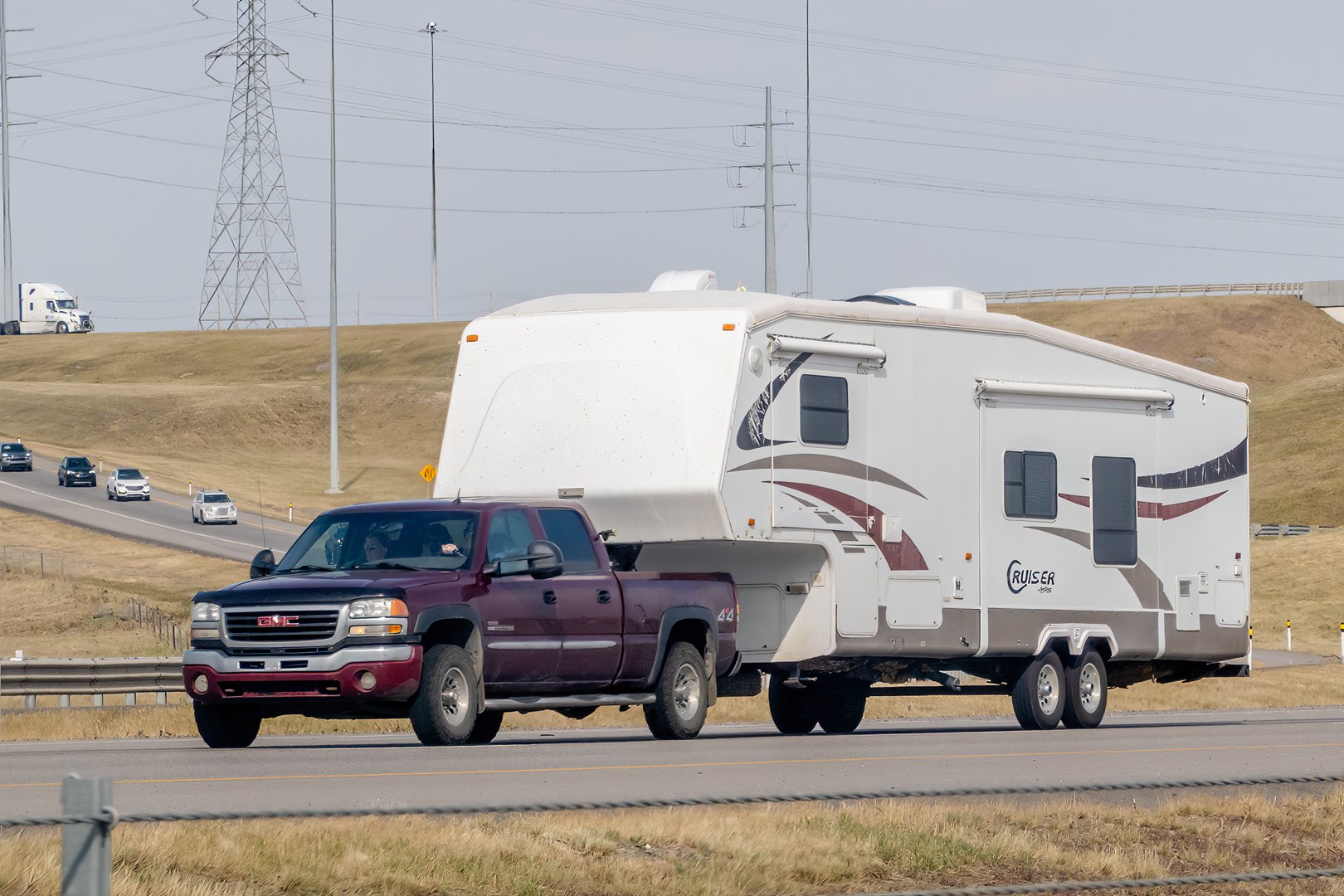 Calgary, Alberta, Canada. Aug 12, 2024. A maroon GMC Sierra HD truck towing a white fifth-wheel RV labeled Cruiser on a multi-lane highway against a backdrop of grassy hills and power lines.