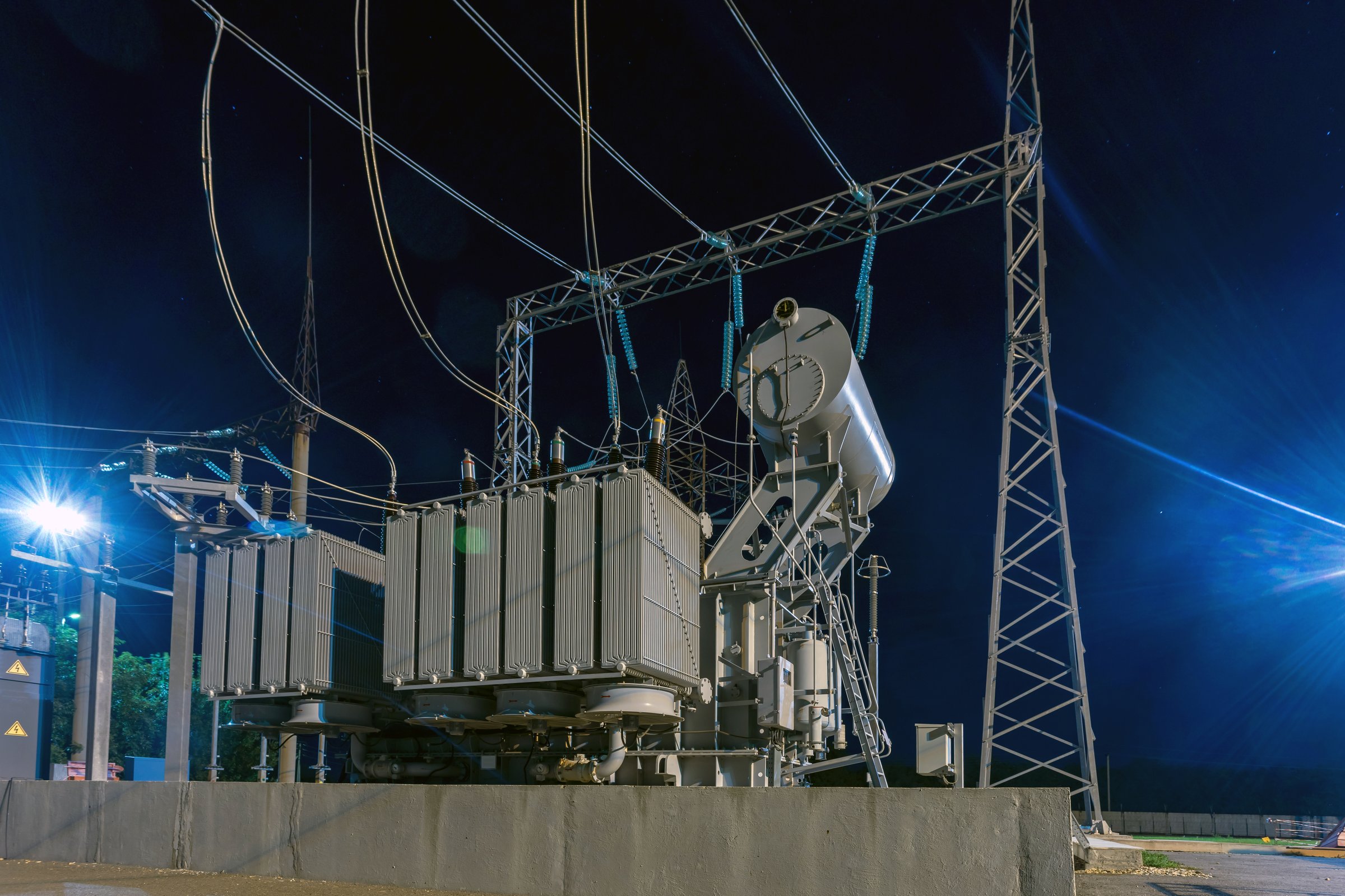 High voltage electrical transformer at power distribution station at night with industrial lighting.