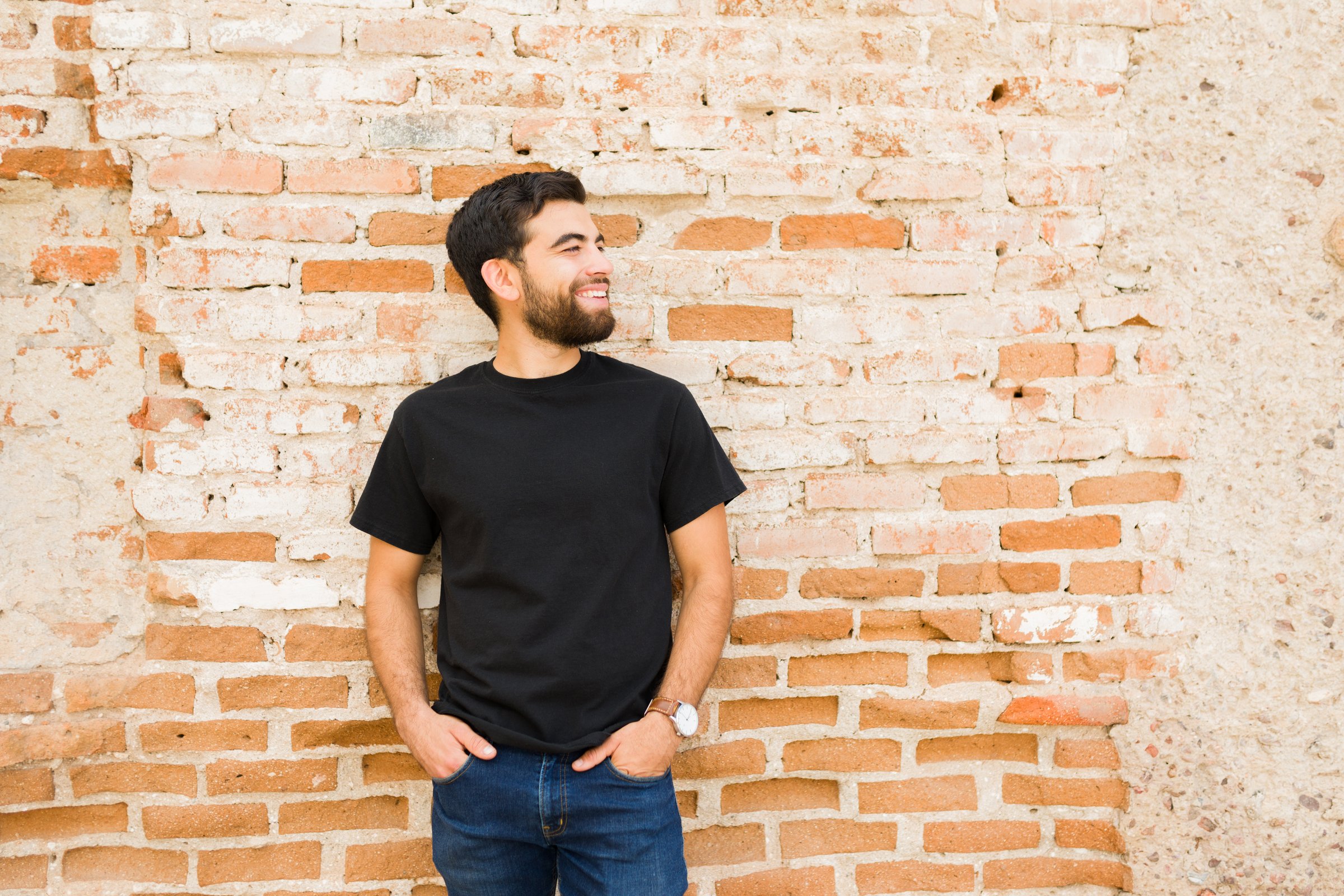 Casually dressed young hispanic man in a plain black t-shirt stands against a textured brick wall, ideal for mock-up designs with a smile on his face