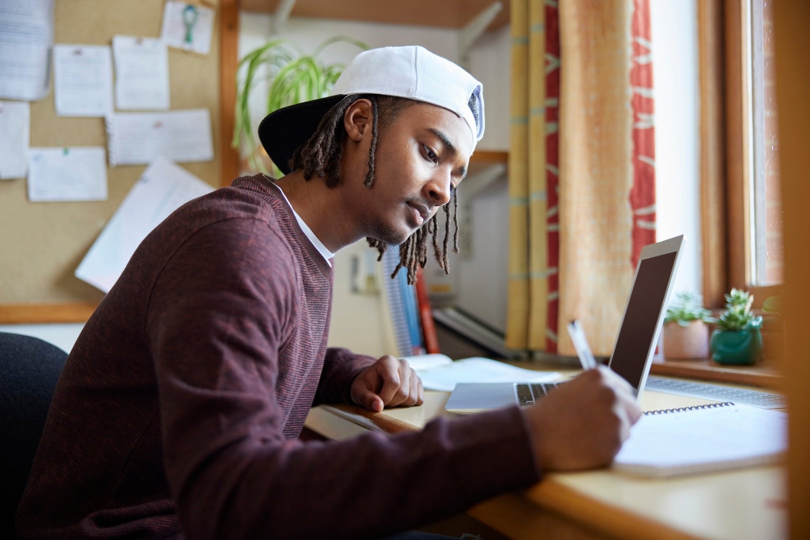 Male University Or College Student Wearing Baseball Cap Studying With Laptop At Desk In Room