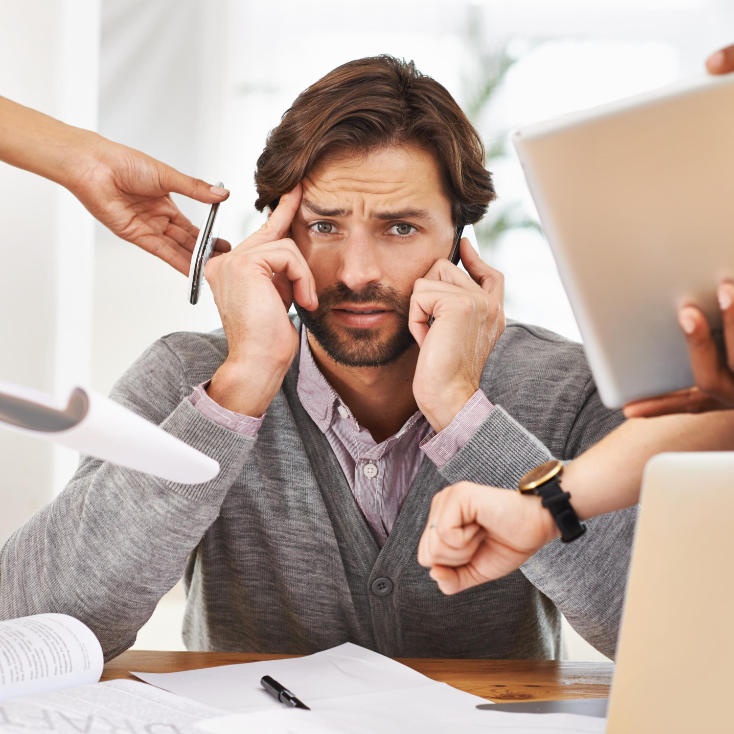 Stressed man at desk surrounded by hands holding a phone, pen, paper, and laptop, symbolizing multitasking pressure.