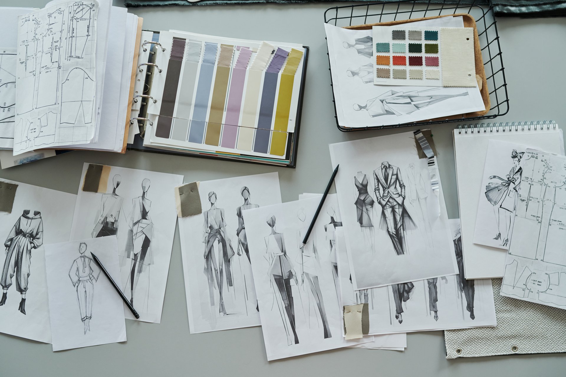 Top view of variety of drawn sketches of new trendy attire and sewing patterns, basket with textile samples and catalogue of swatches