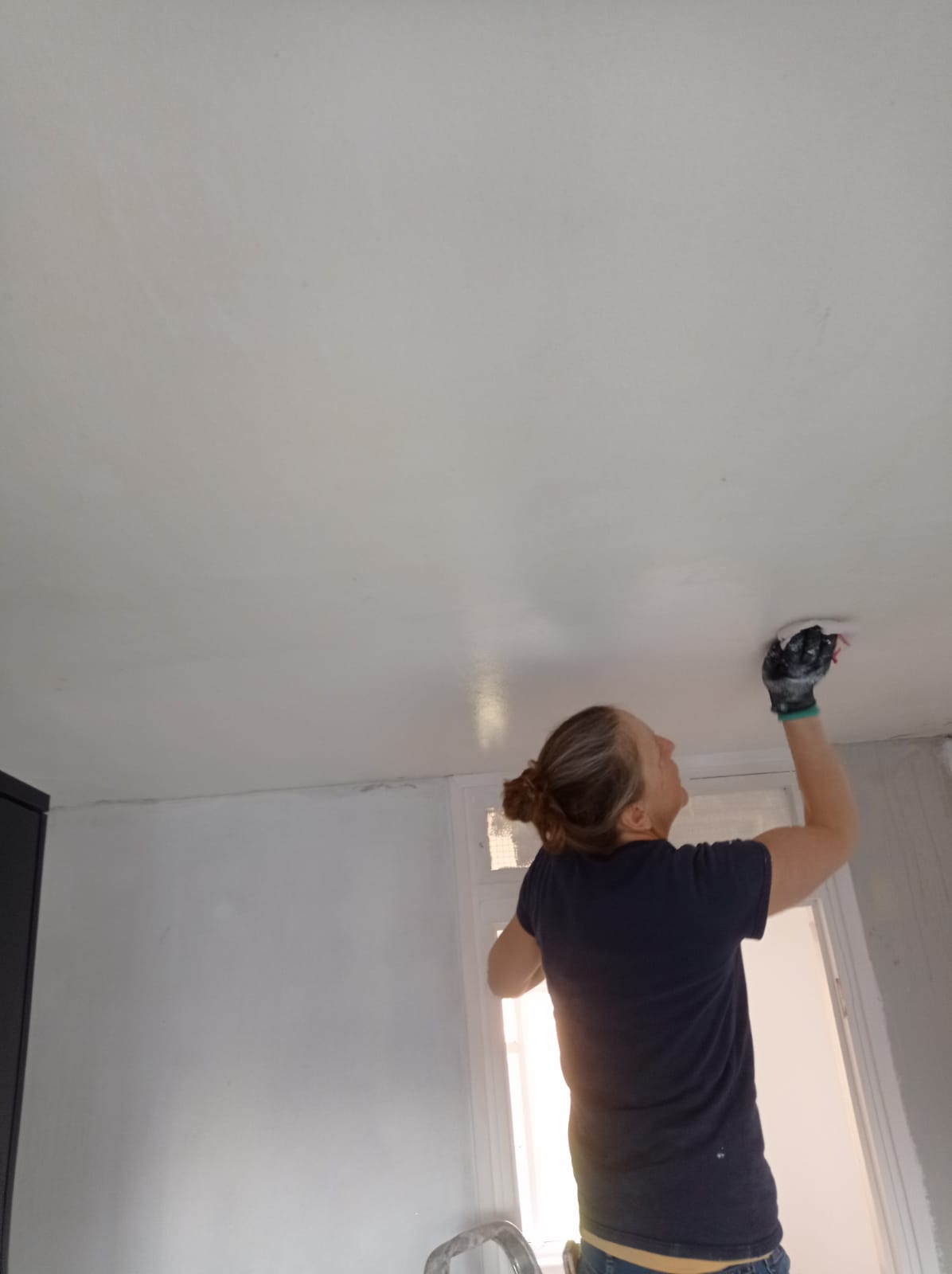 Person standing on a ladder, painting a white ceiling with a roller in a room with natural light.
