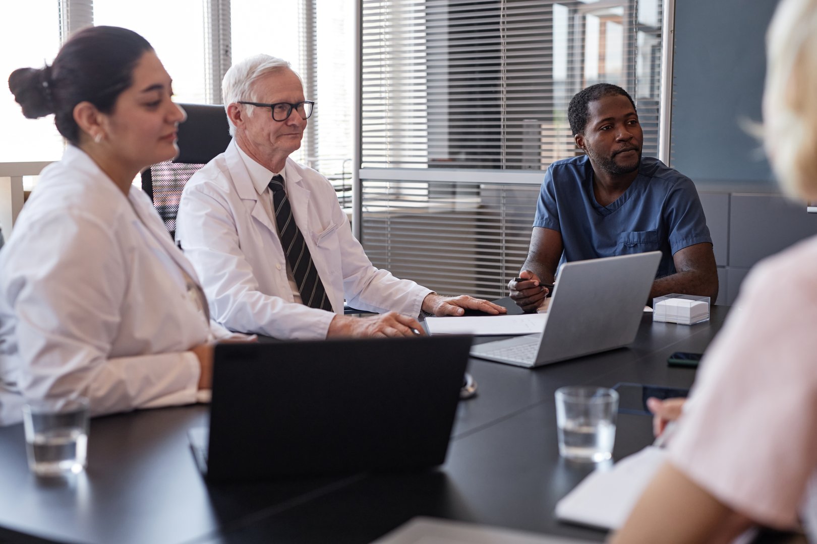 African American male clinician in blue scrubs engaged in group discussion with colleagues about anamnesis of patients disease, while sitting at meeting table at medical office