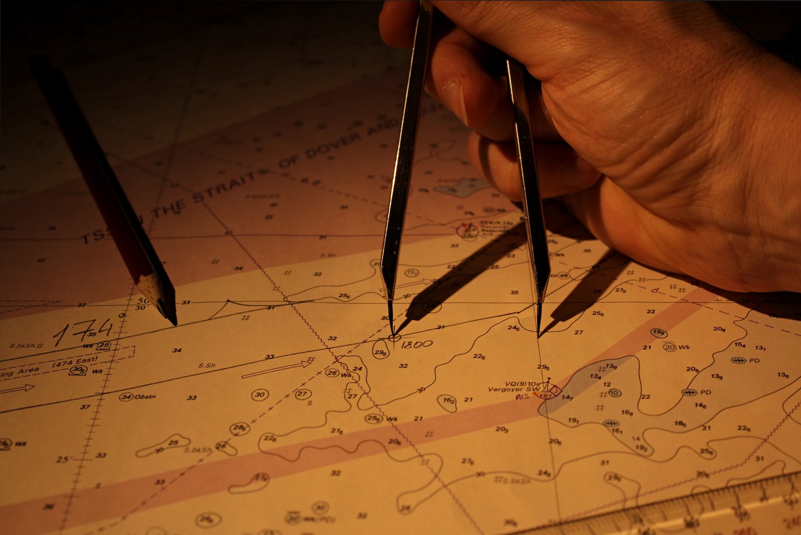 A close-up of a hand using dividers to plot the position on a nautical chart, illuminated by warm light. The chart features depth soundings and navigational information, suggesting the planning of a sea voyage or maritime navigation.  A pencil and ruler are also visible, emphasizing the traditional tools and methods of charting a course.