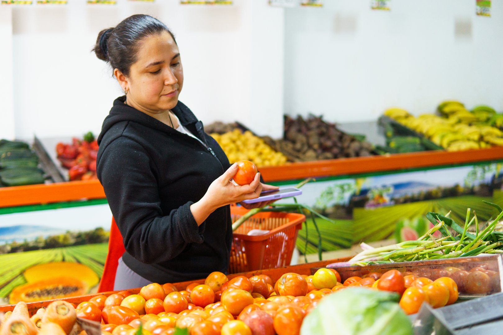 A female domestic worker is shopping for lunch at a fruit and vegetable store. He is looking at a tomato and has a market basket in his arm