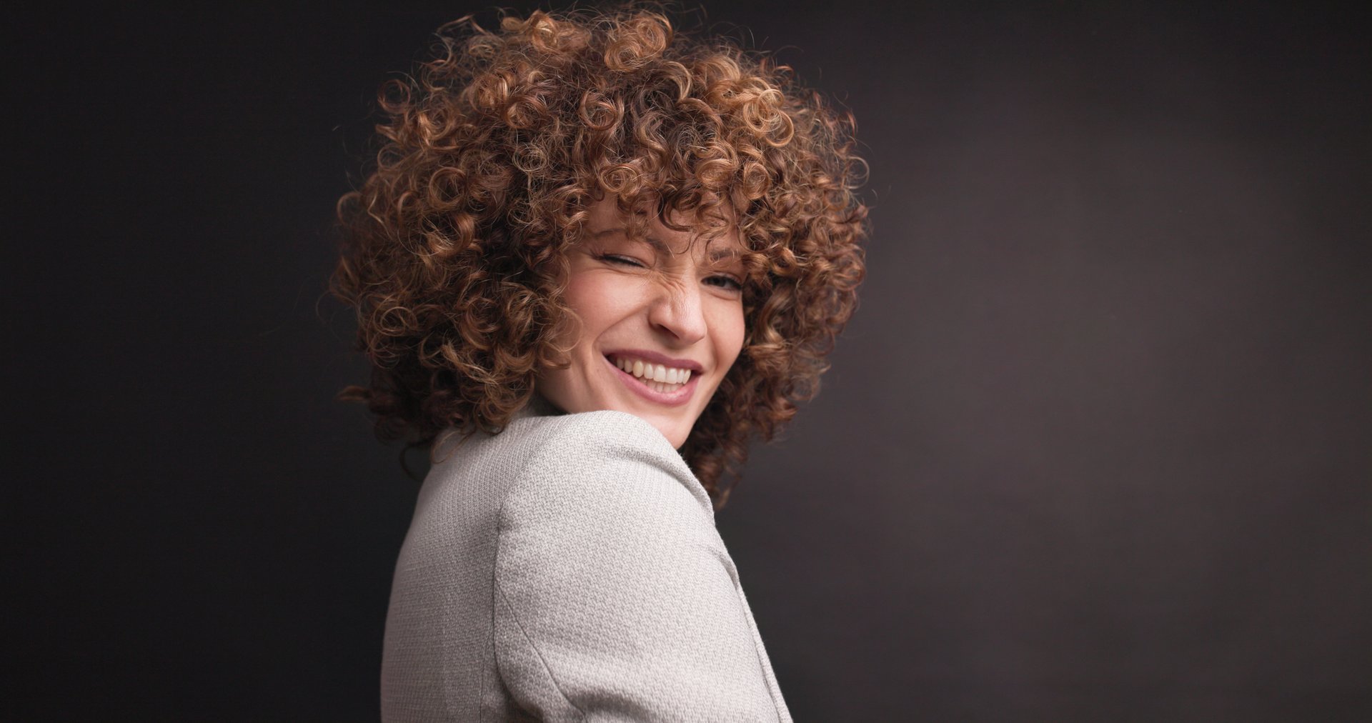 Portrait of smiling young businesswoman with short curly hair in stylish blazer looking at camera against black background