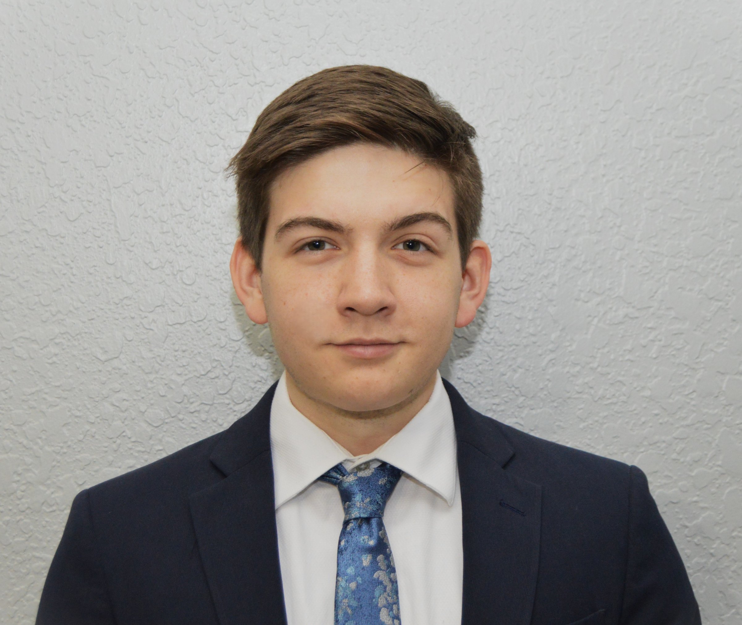 Young man in a suit and tie stands against a textured light gray wall, looking directly at the camera.