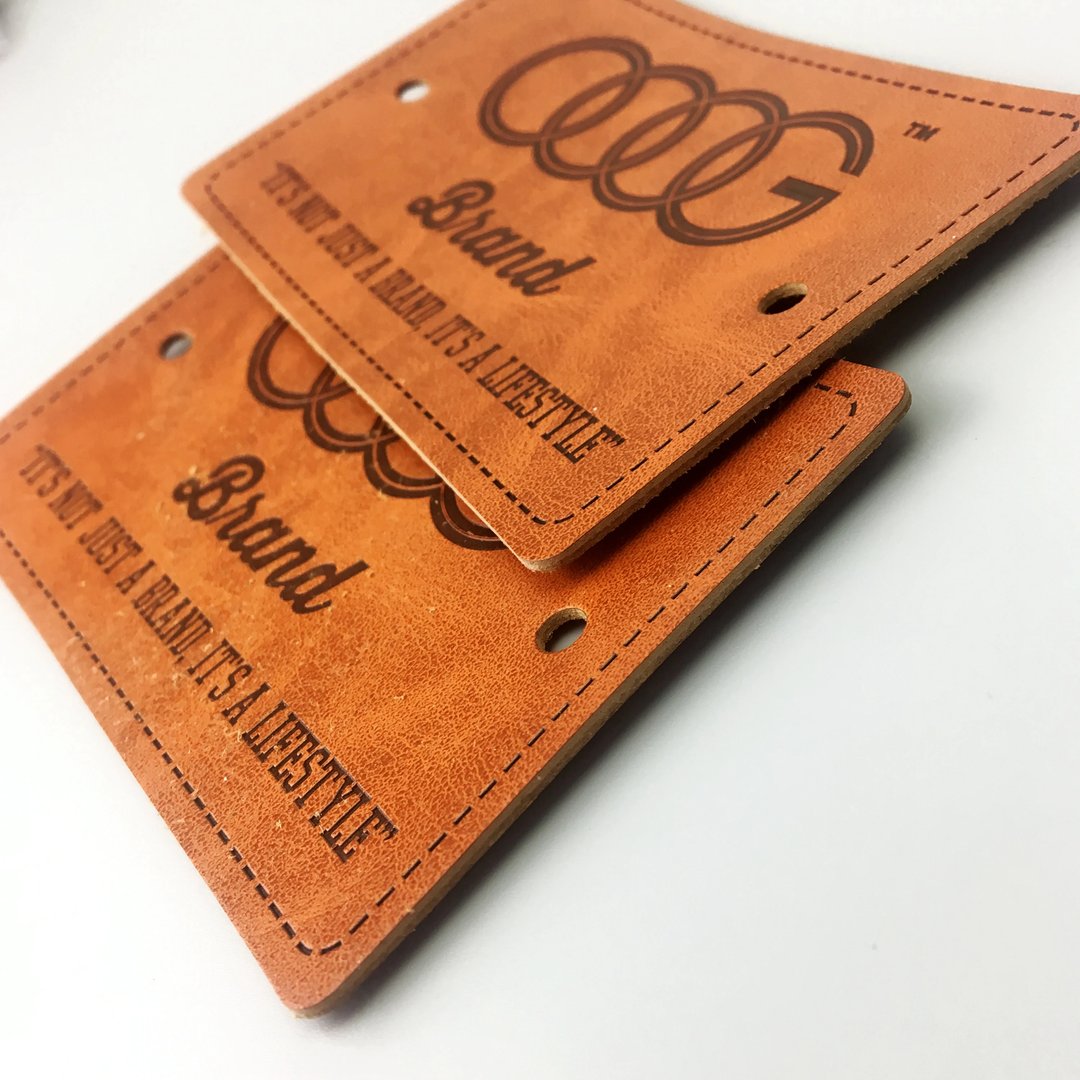 Two leather tags with embossed text and round holes, displaying a brand logo and slogan on a white surface.