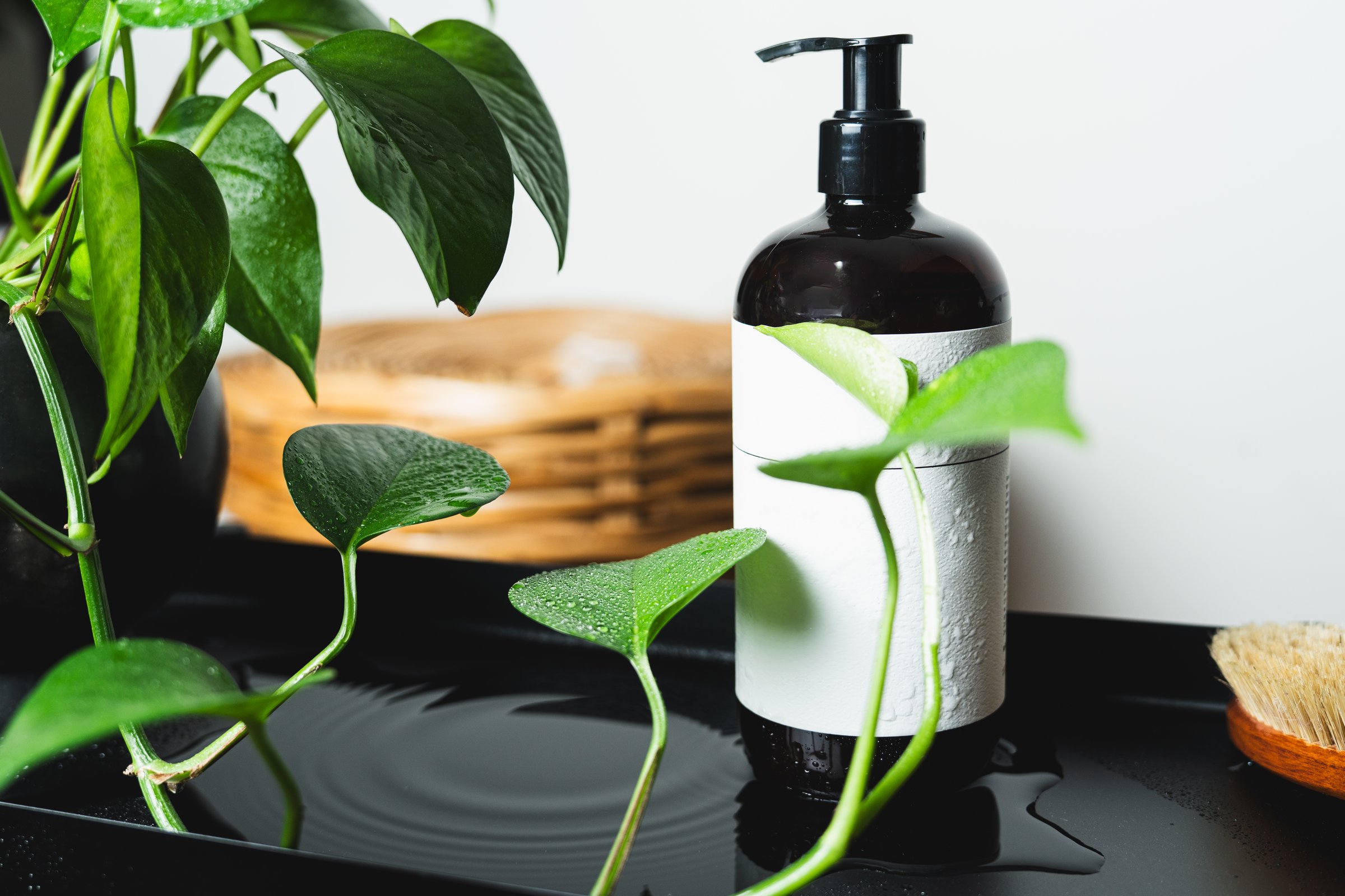 Dark amber dispenser bottle with blank white label, fresh green leaves and water drops on black tray