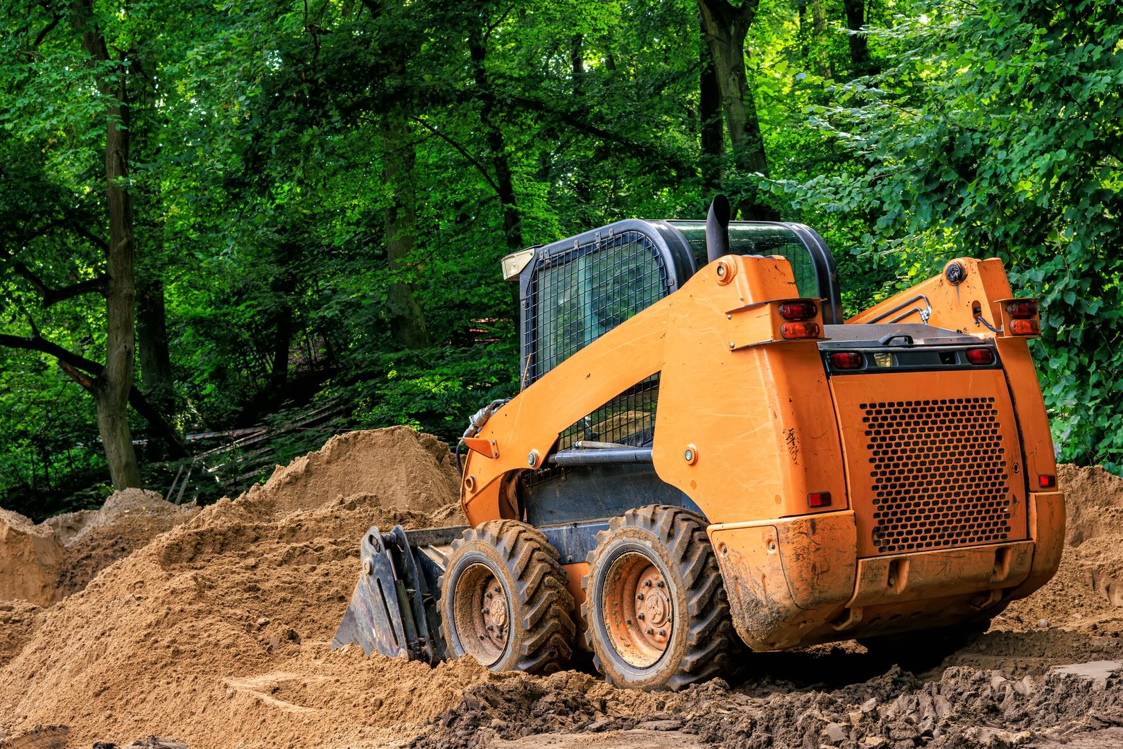 An orange skid steer loader shovels soil in a park.