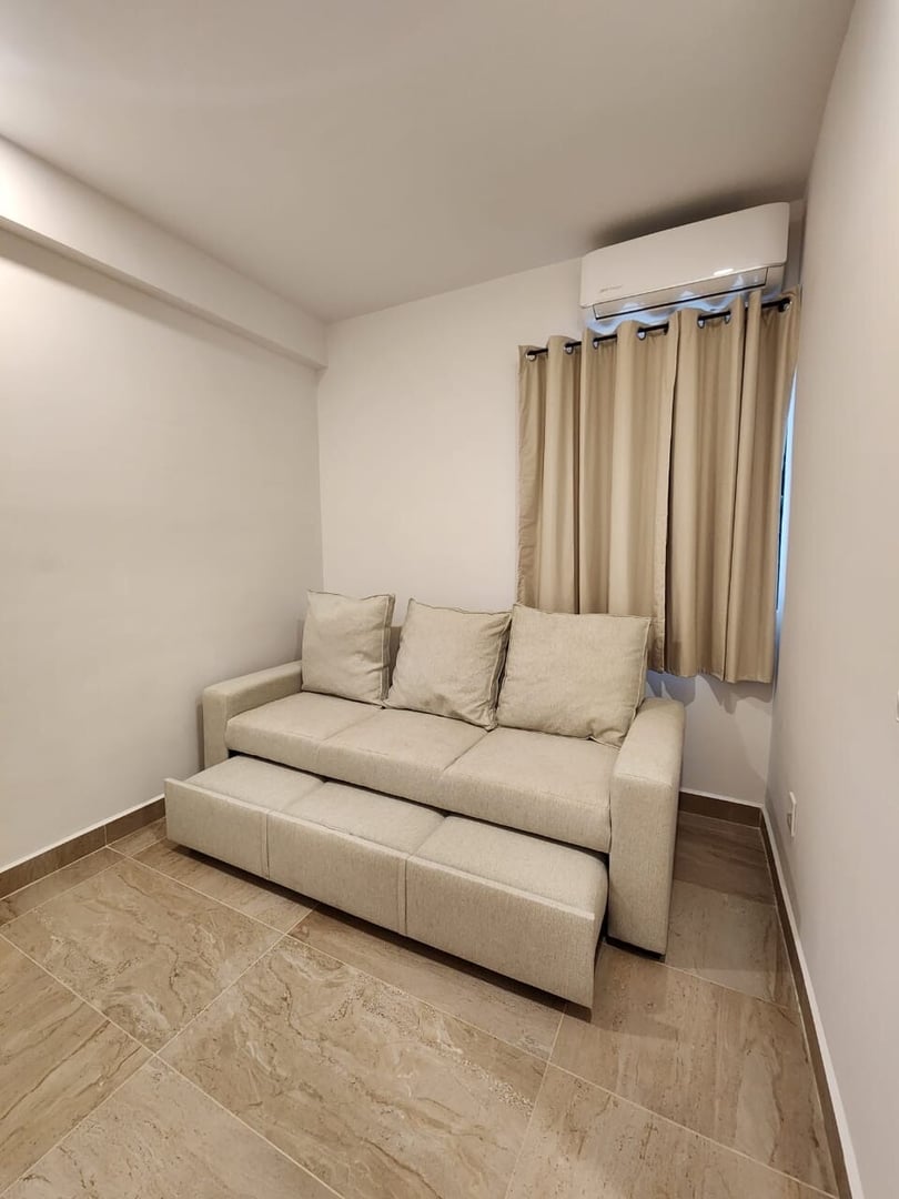 Small beige sofa with pull-out bed in a simple room with tiled floor, beige curtains, and wall-mounted air conditioner.
