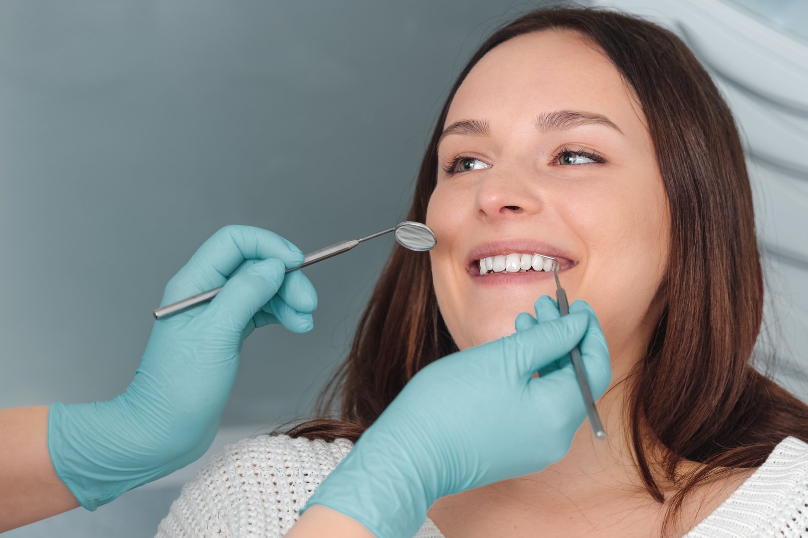 Portrait of happy young woman with open mouth sitting in dental chair, dentist holding in hands in blue gloves mouth mirror and periodontal explorer scaler and checking caries, healthy teeth concept