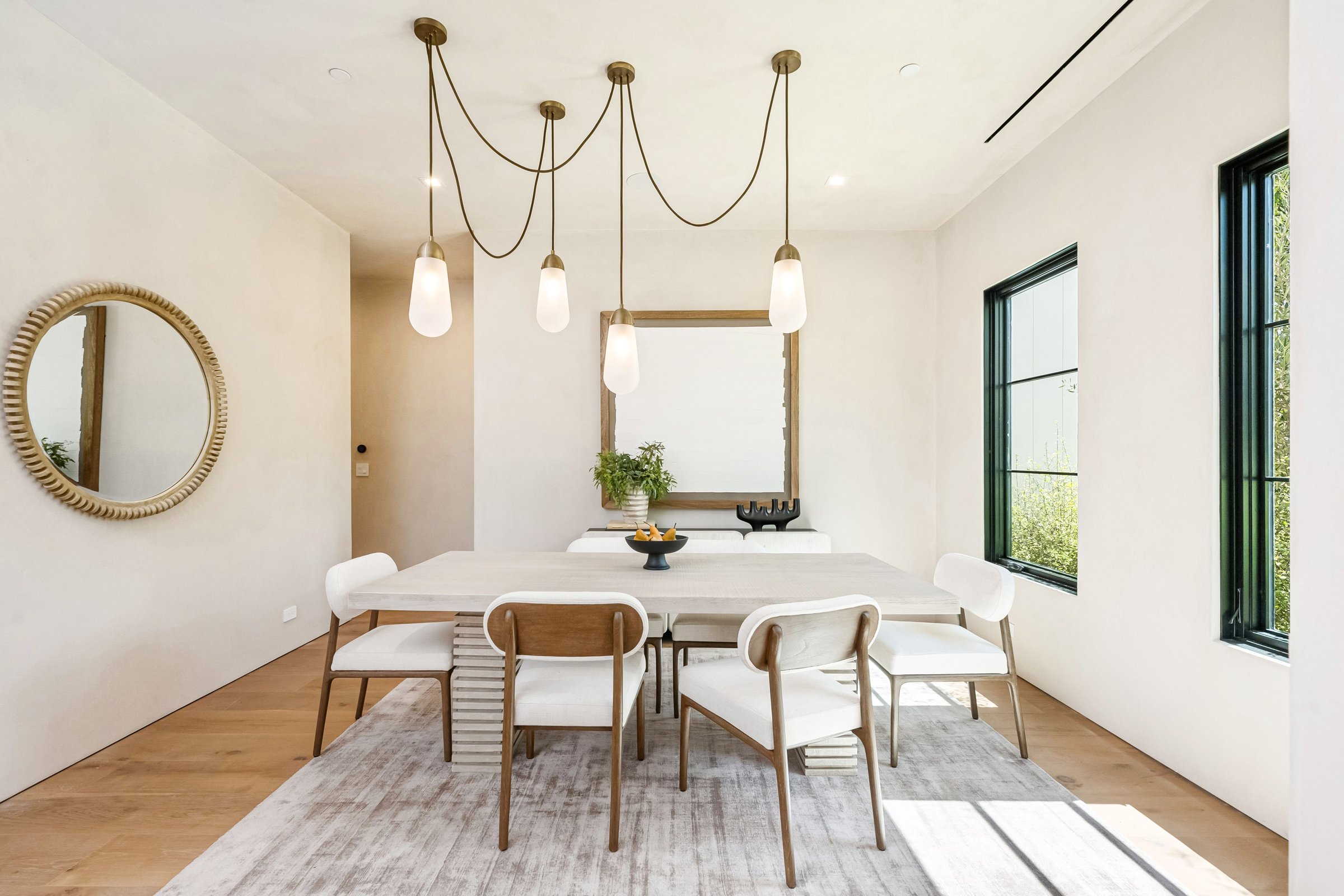 Modern dining room with elegant pendant lights, wooden table, and large mirror, featuring natural light and minimalist decor.