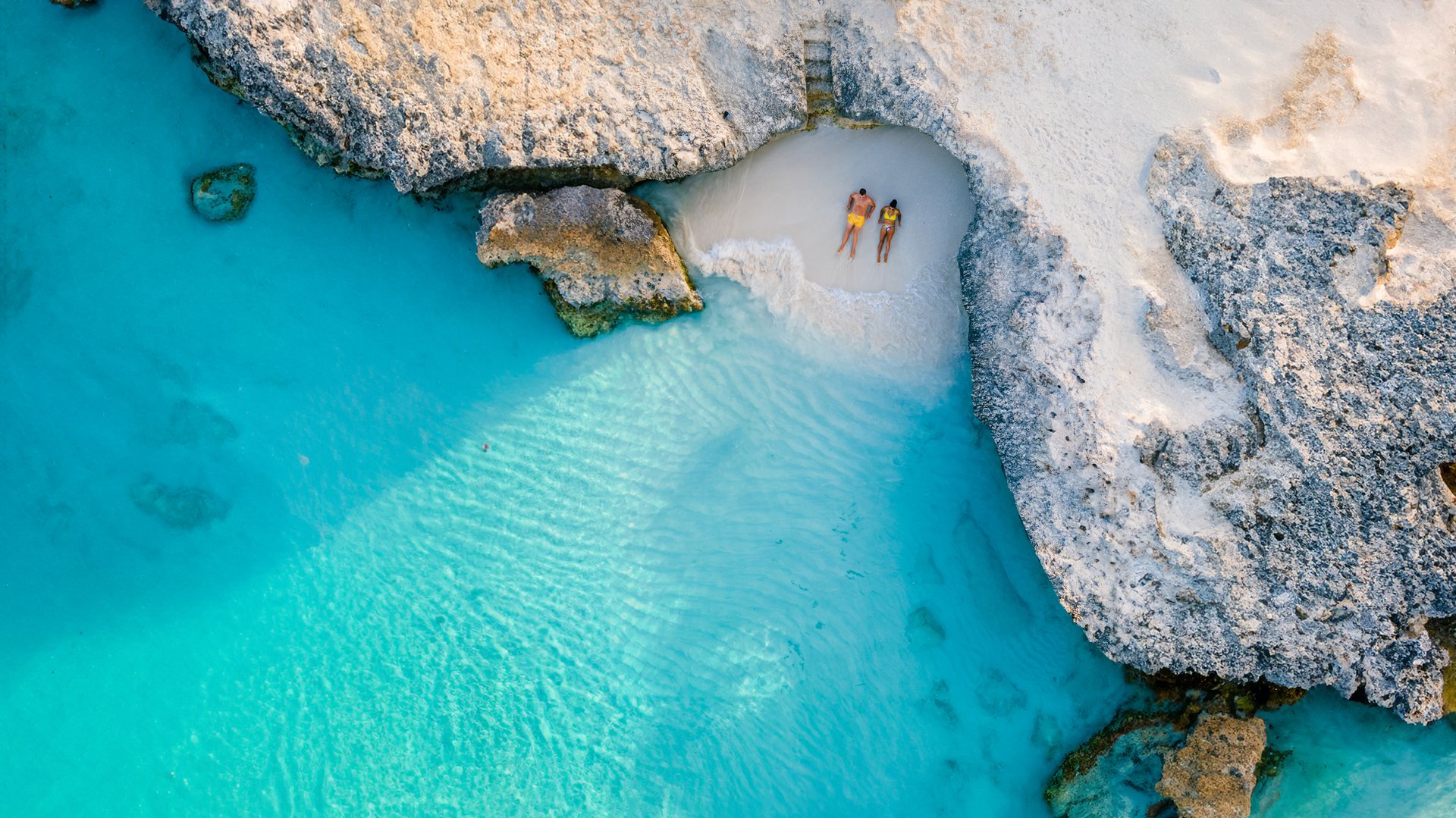 Aerial view of two people sunbathing on a small sandy beach surrounded by turquoise water and rocky cliffs.
