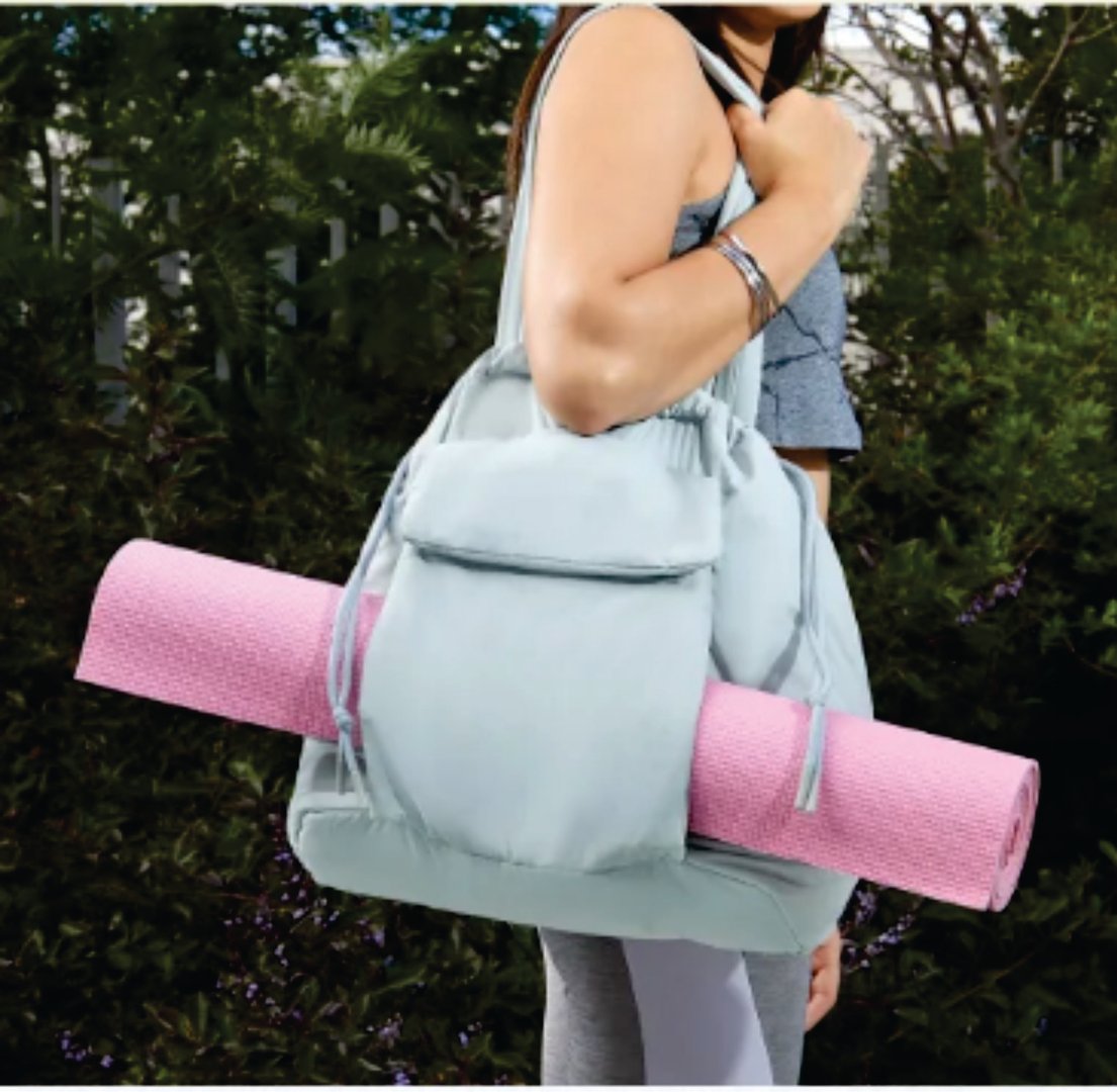 Woman carrying a yoga mat secured in a light blue shoulder bag, outdoors. She wears a tank top and leggings.