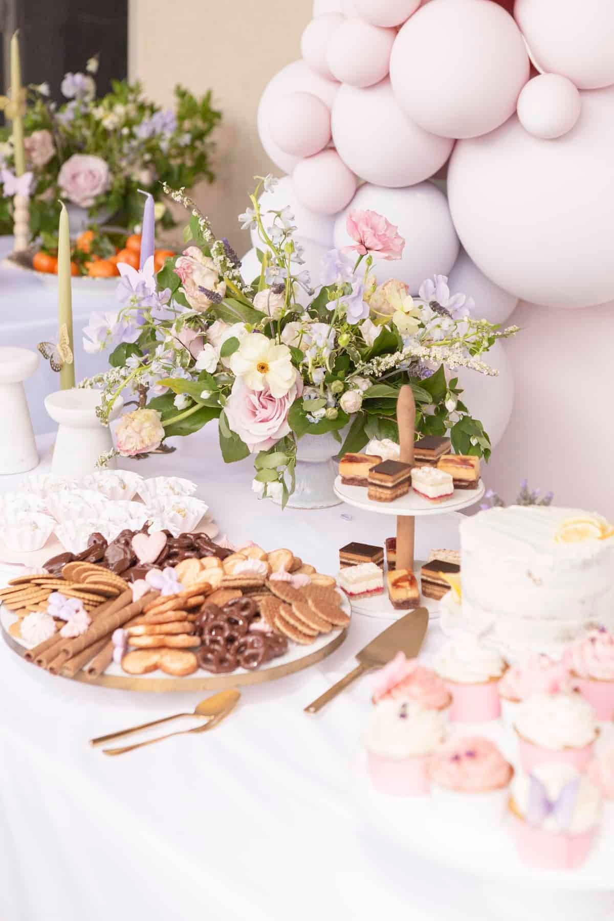 Dessert table with pastries, cupcakes, a cake, and floral arrangements, decorated with pastel balloons and candles.