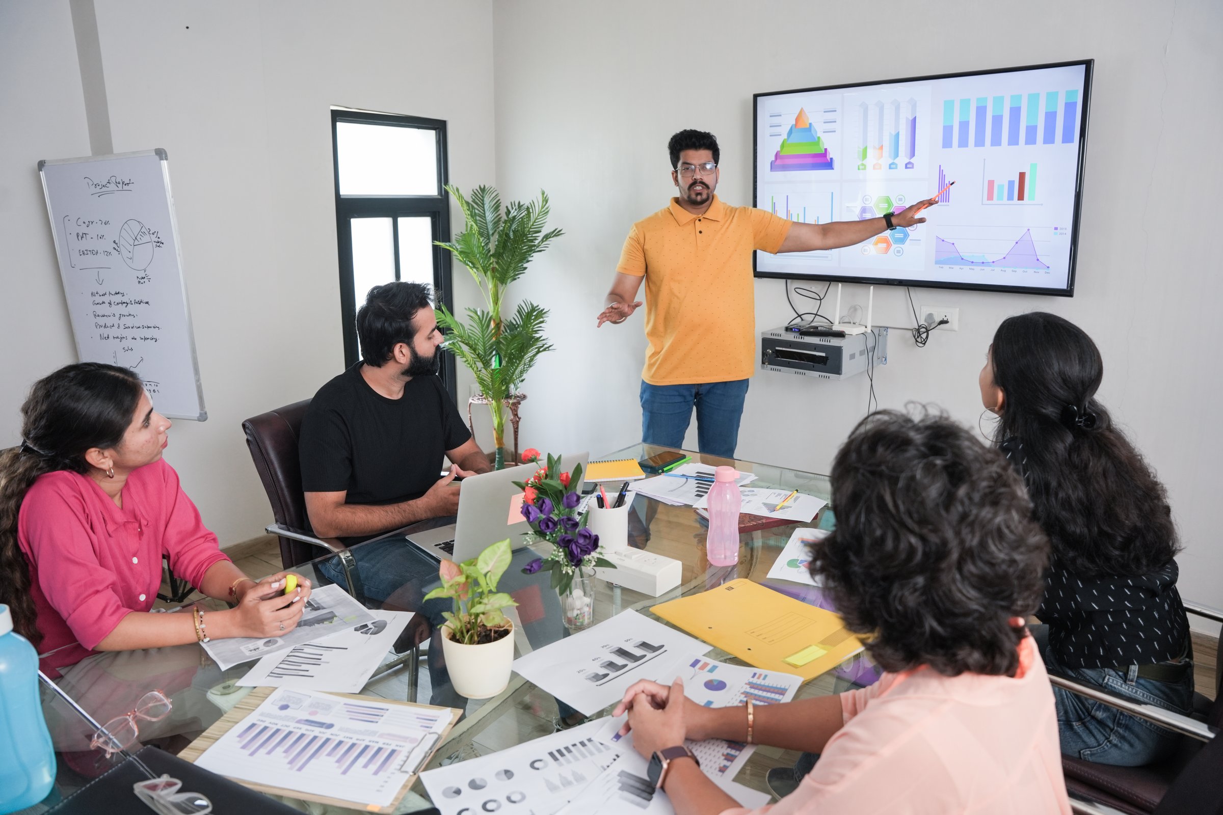 young indian man entrepreneur presents financial data with charts and graphs on wall tv in the business meeting room to colleagues for a start-up project.