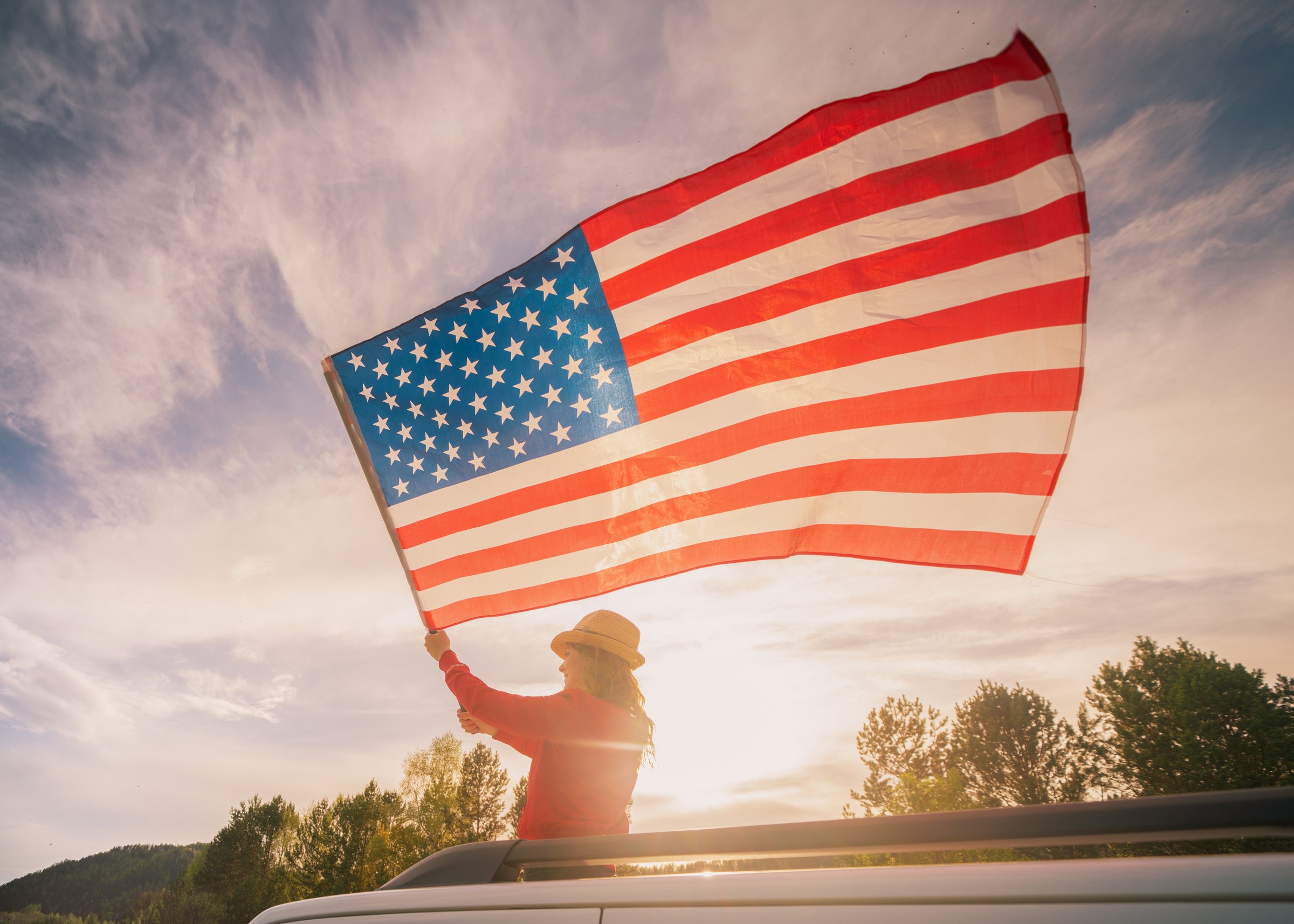 Close up on USA flag in a hand of a person against the sky. Independence Day or traveling in America concept, A woman waves a flag from a car hatch.